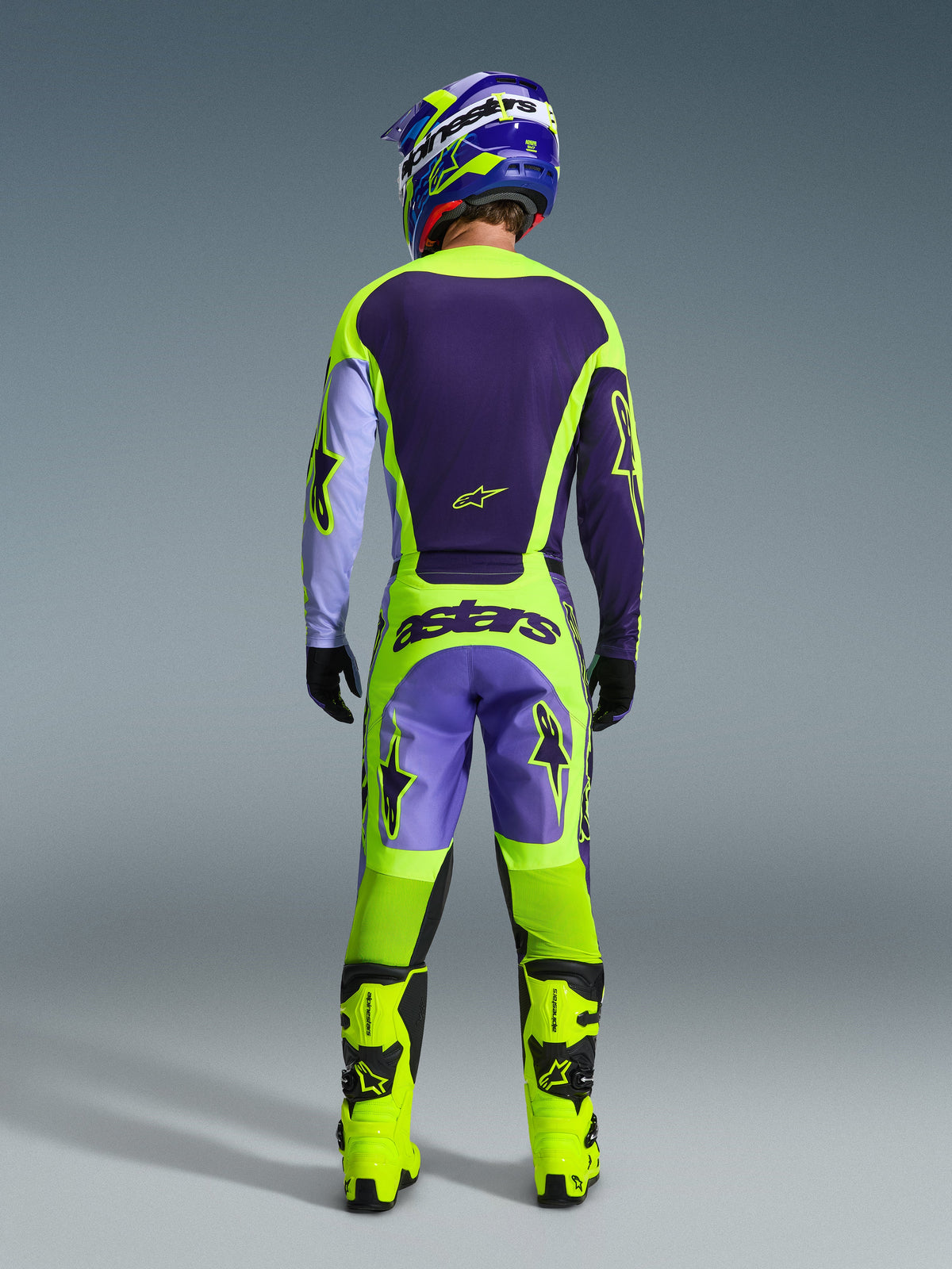 Motocross-Fahrer im Alpinestars 2026 Racer Portl Ausrüstungsset, Rückansicht, dunkellila und neongelbes Jersey mit lavendelfarbenen Ärmeln, passende lila und neongelbe Hose mit 'astars'-Branding, leuchtend neongelbe Stiefel, professionelles Offroad-Rennoutfit