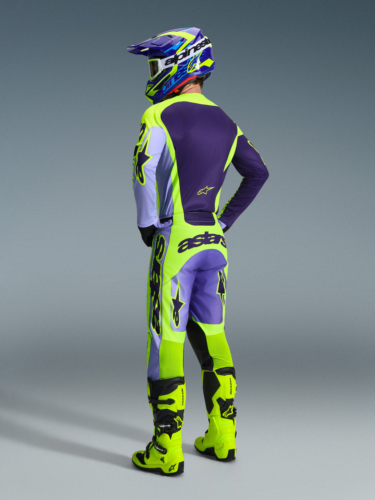 Motocross-Fahrer im Alpinestars 2026 Racer Portl Gear Set, Jersey und Hose in Lila und Neongelb mit lavendelfarbenen Einsätzen, passender lila-gelber Helm, leuchtend neongelbe Stiefel, Rückansicht zeigt professionelle Offroad-Rennausrüstung.