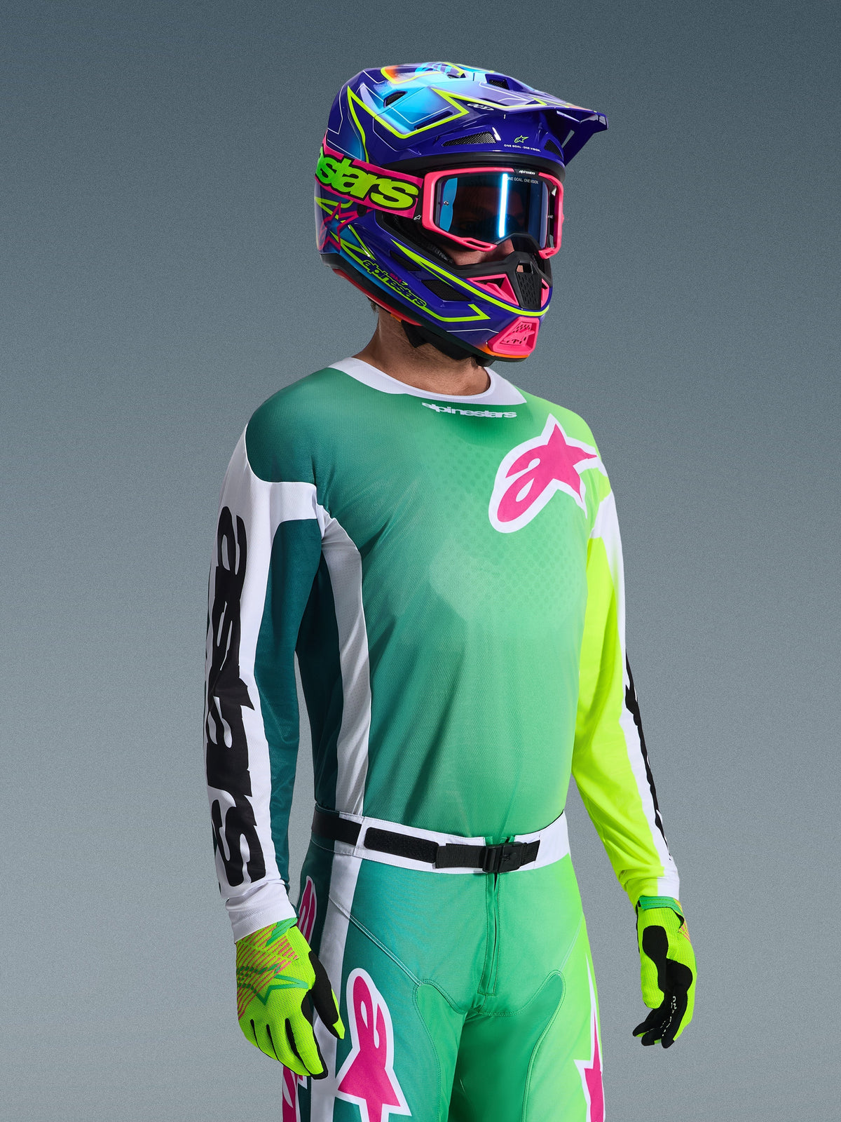 Alpinestars 2026 Racer Portl Trikot, Motocross-Trikot, Grün, Weiß und Schwarz, getragen von einem Fahrer mit passender grüner Hose, fluoreszierenden Handschuhen und einem leuchtend blau-violetten Helm mit pinkfarbener Brille, mit einem pinken Astars-Logo auf der Brust.