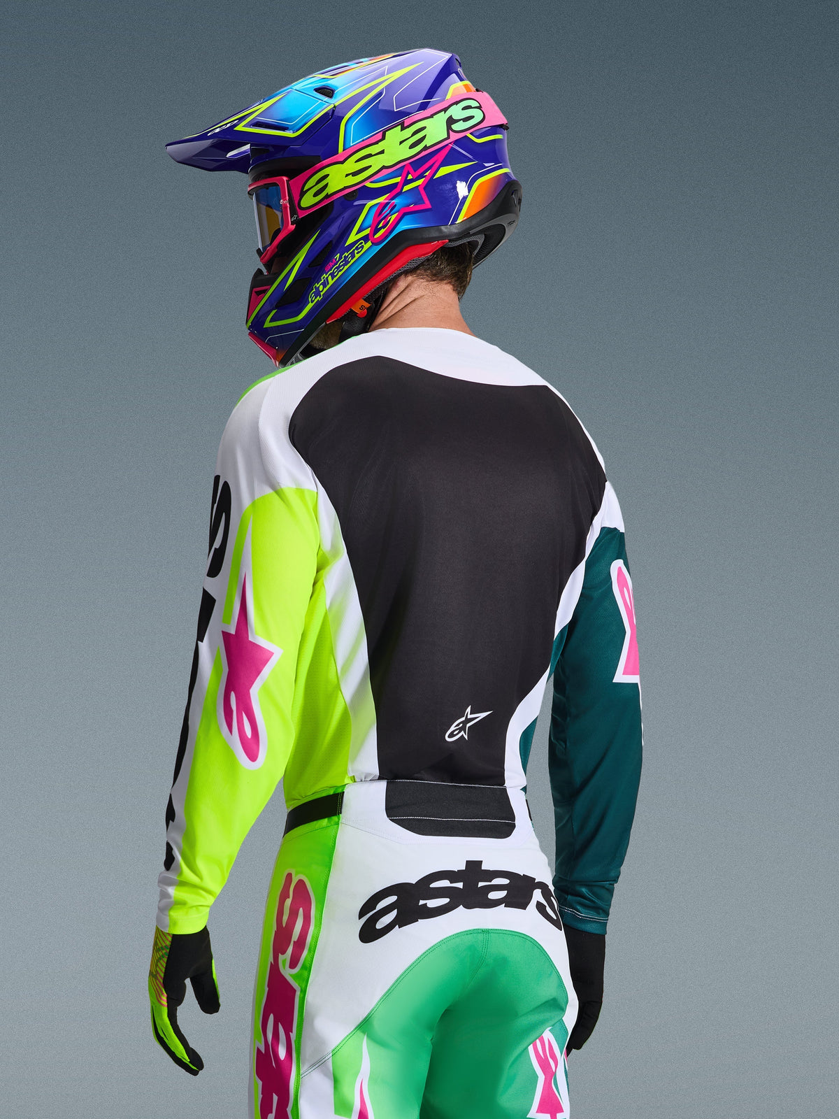 Motocross-Fahrer in der Rückansicht mit dem Alpinestars 2026 Racer Portl Trikot in Grün-Weiß-Schwarz, rosa Stern-Logos, passender Hose und einem bunten blau-violetten Helm, koordinierte Rennausrüstung für Offroad-Performance.