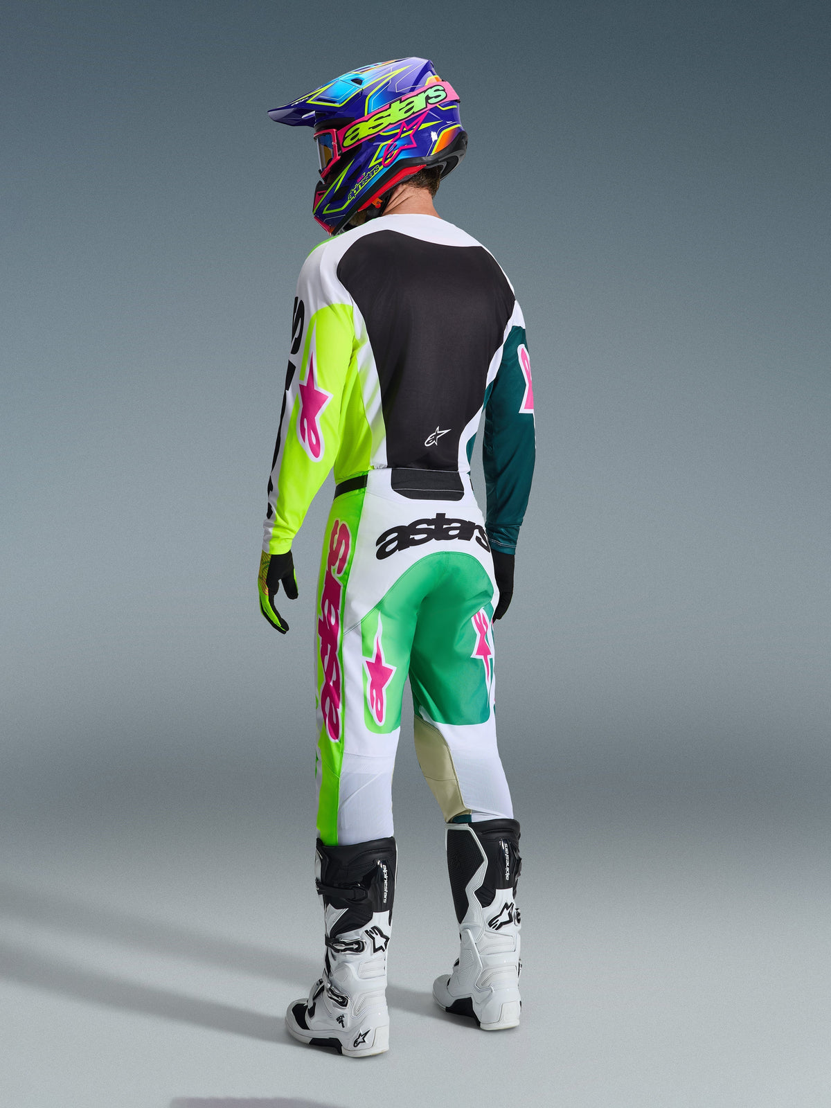 Motocross-Fahrer im Alpinestars 2026 Racer Portl Trikot in Grün Weiß Schwarz, von hinten gesehen, passende Rennhose mit pinkfarbenen Stern-Logos und „astars“-Branding, weiße und schwarze Stiefel und ein bunter Helm, komplettes abgestimmtes Outfit für den Offroad-Rennsport