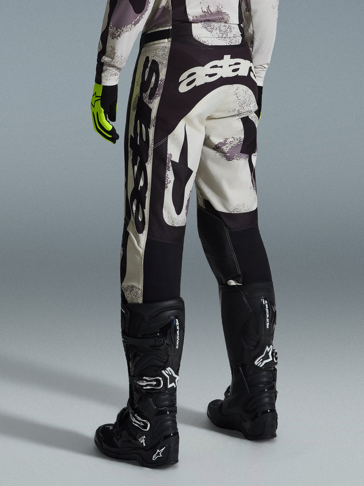 Alpinestars 2026 Racer Lahnd Hose, Motocross-Hose, Sand-Camo und Grau, Rückansicht mit markantem weißen Branding an Gesäß und Beinen, kombiniert mit schwarzen Motocross-Stiefeln und gelben Handschuhen, professionelle Offroad-Rennausrüstung