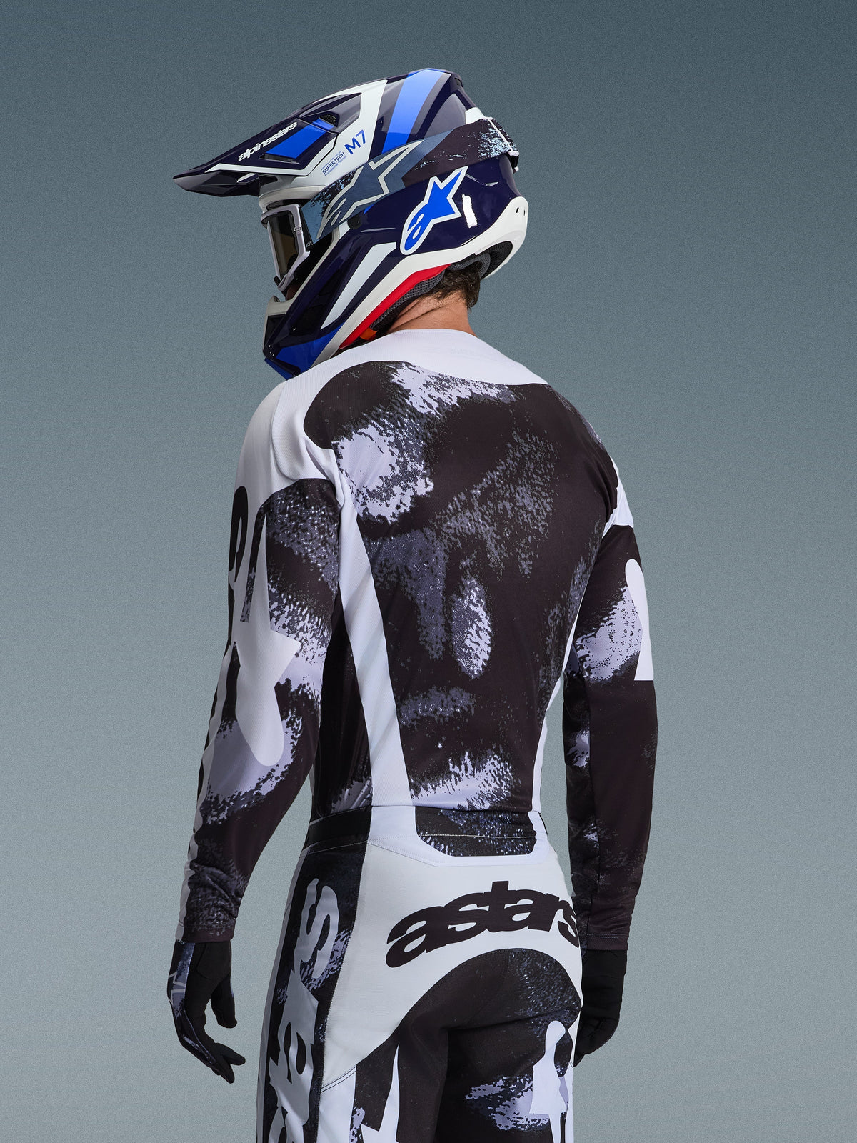 Alpinestars 2026 Racer Lahnd Jersey in Iron Camo, Rückansicht eines Motocross-Fahrers in einer Ausrüstung mit schwarz-weiß-grauem abstraktem Muster, inklusive passender Hose, schwarzen Handschuhen und einem blauen Rennhelm mit Brille