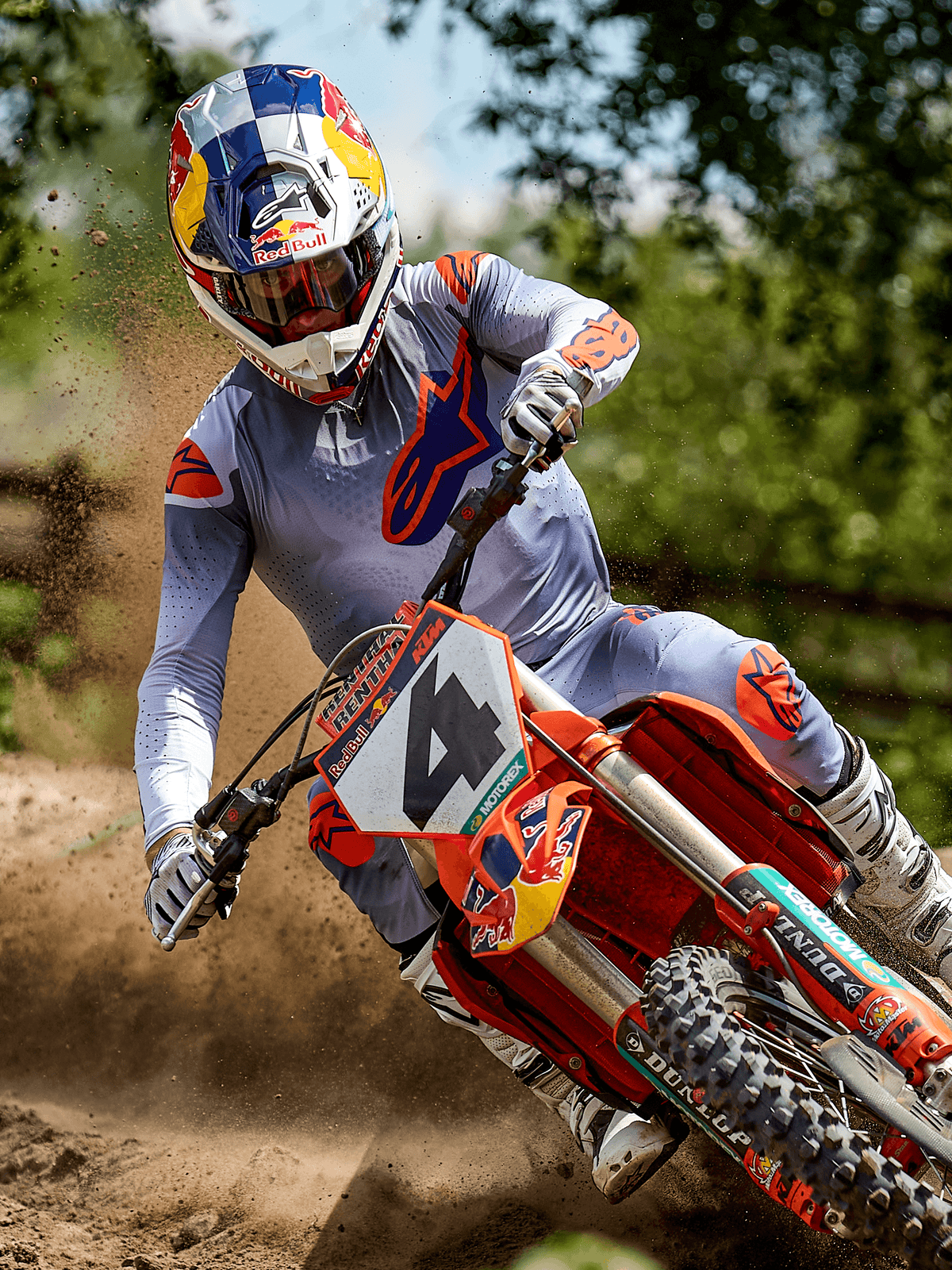 Motocross-Fahrer in Aktion auf einer Cross-Strecke im Alpinestars 2025 Supertech Lipan Jersey in Grau Orange Fluo, passende graue Hose mit orangefarbenen Akzenten, weiße Stiefel und Red Bull-Helm, Kurvenfahrt auf einem orangefarbenen Motocross-Bike mit aufwirbelnder Erde, professionelle Offroad-Rennausrüstung.