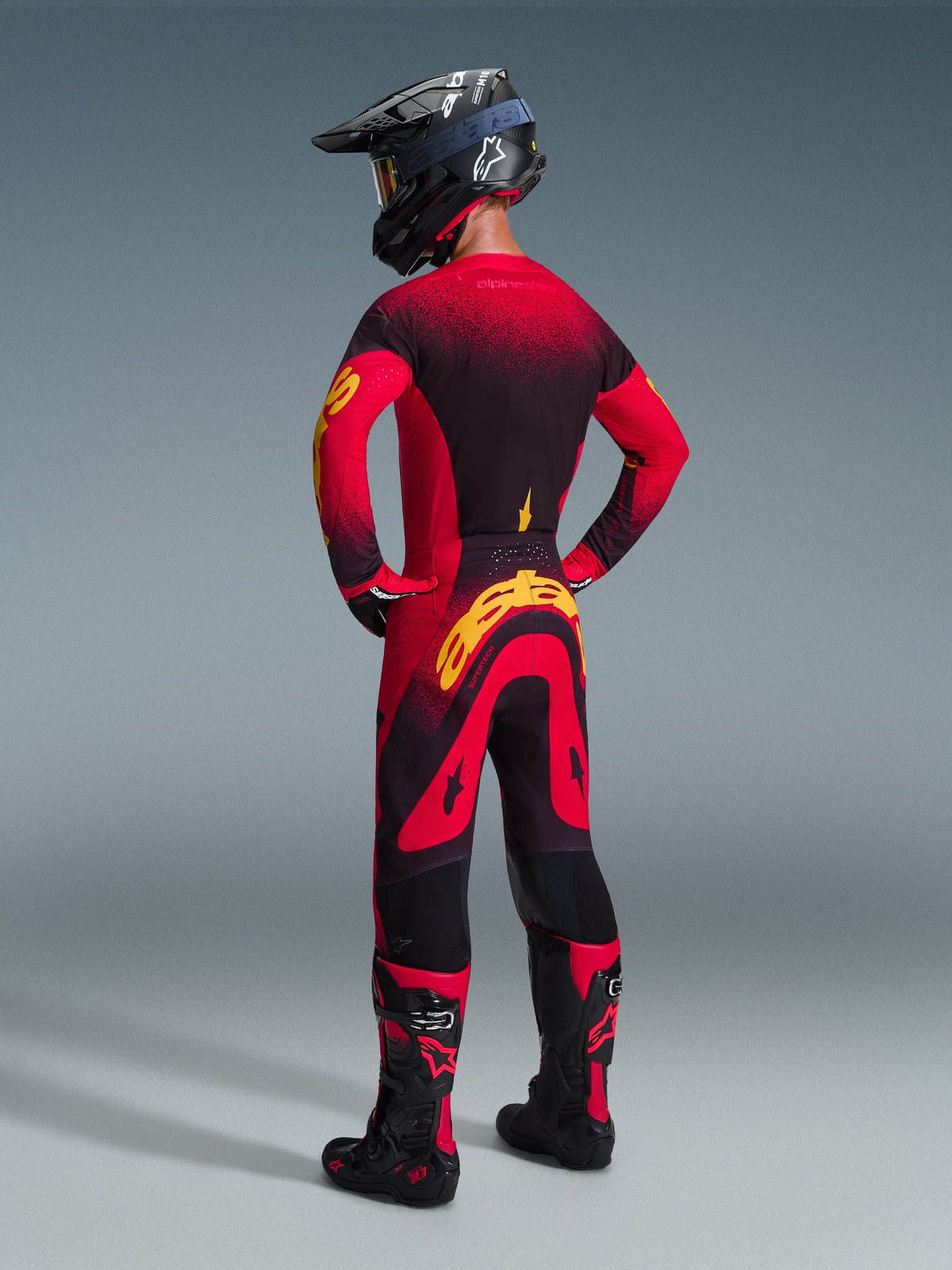 Motocross-Fahrer in kompletter Rennausrüstung von hinten gesehen, Alpinestars 2026 Supertech Scenz Trikot in Rot, Schwarz und Gelb, passende Motocross-Hose mit gelbem Branding, schwarzer Helm und schwarz-rote Stiefel, abgestimmtes Outfit für professionellen Offroad-Rennsport