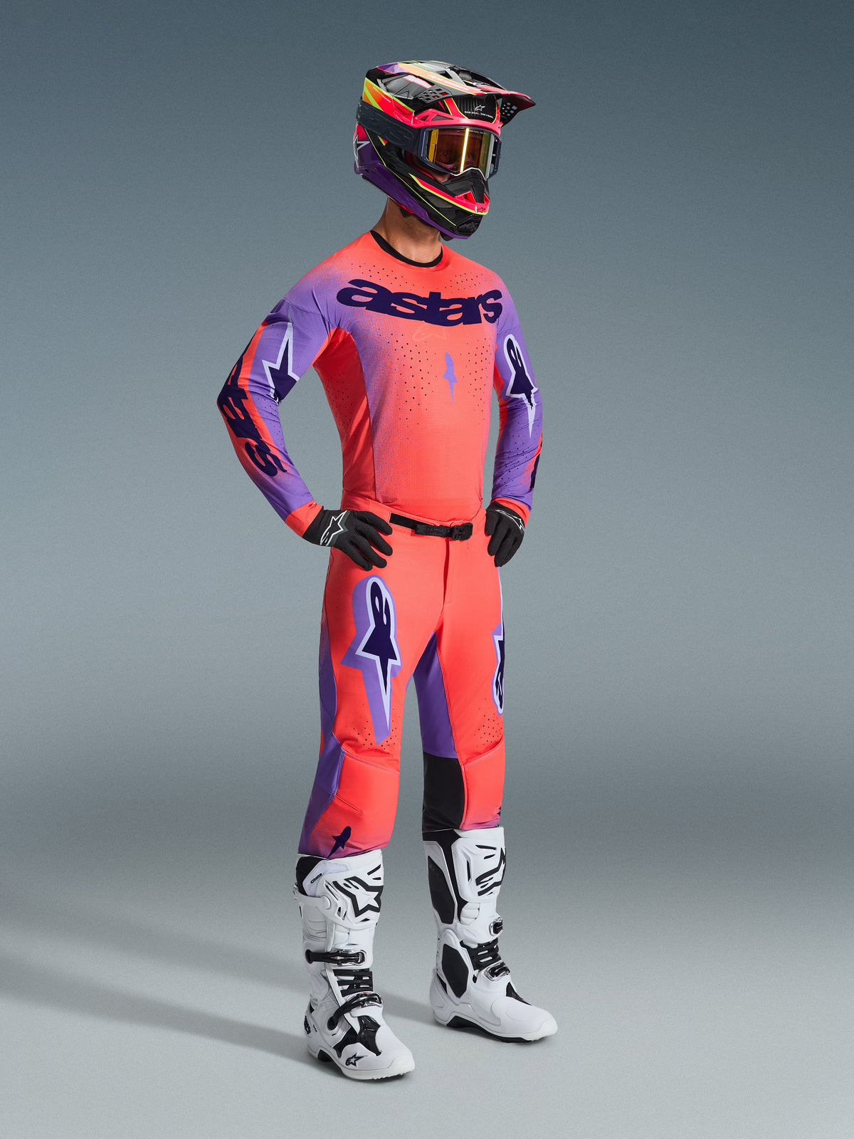 Motocross-Fahrer im kompletten Alpinestars 2026 Supertech Scenz Gear Set, Trikot in Hot Coral mit lila Ärmeln und 'astars'-Branding, passende korallenrote Hose mit lila Akzenten, mehrfarbiger Helm mit gold getönter Brille, weiß-schwarze Stiefel, schwarze Handschuhe, koordinierte Profi-Rennausrüstung für Offroad-Performance