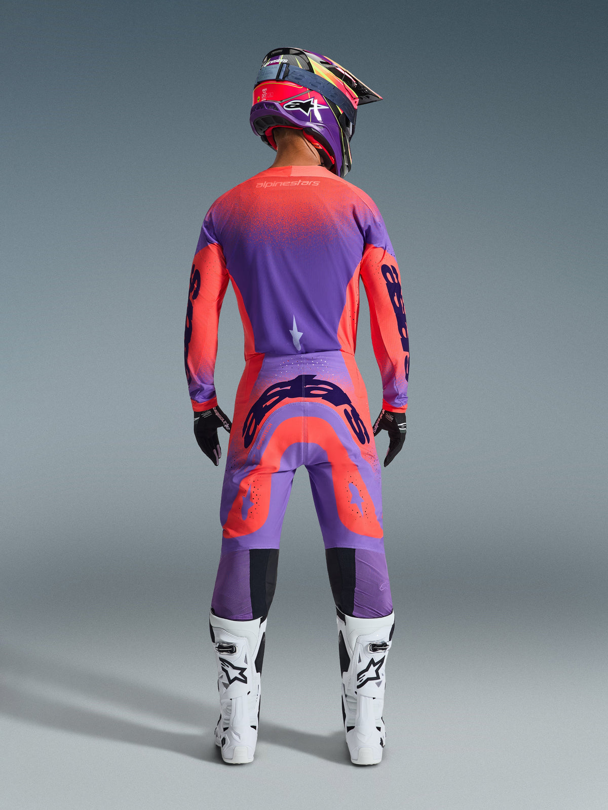 Motocross-Fahrer von hinten im 2026 Supertech Scenz Gear Set in Hot Coral Purple, leuchtendes Trikot und Hose mit Farbverlauf, Alpinestars-Logos, passender Helm, weiß-schwarze Stiefel und schwarze Handschuhe, professionelle Ausrüstung für Elite-Offroad-Rennsport