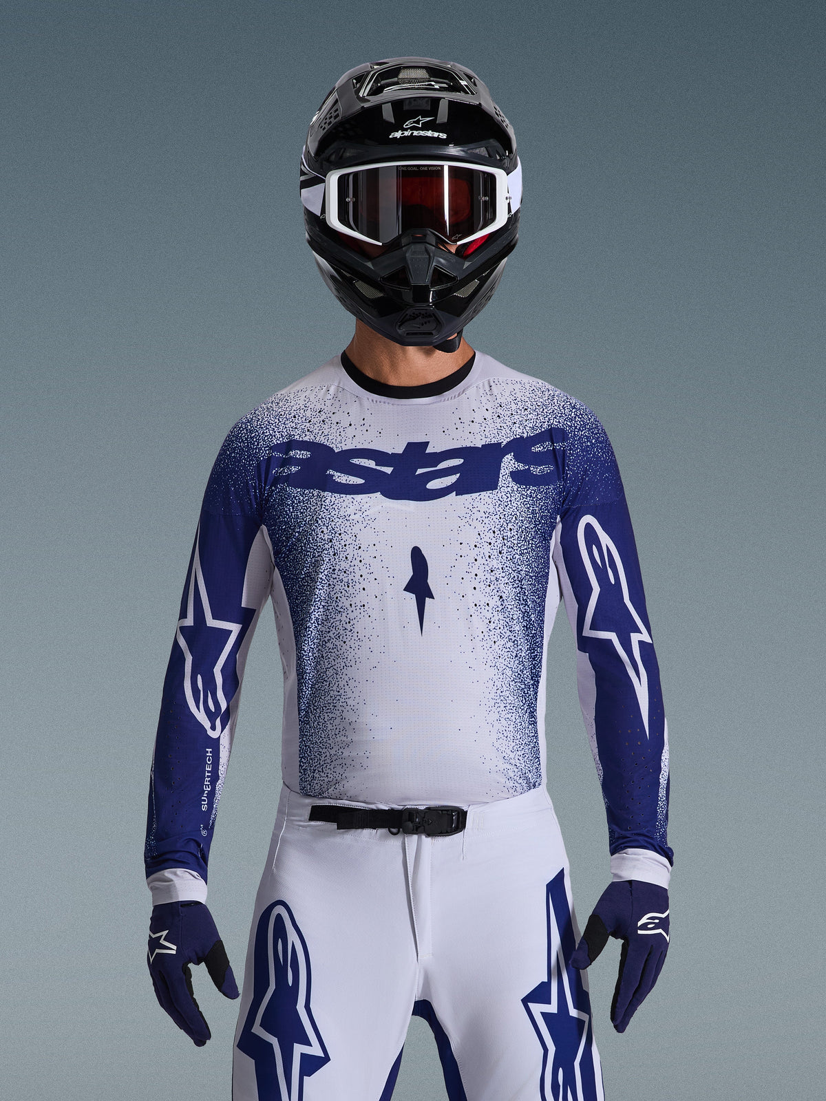 Motocross-Fahrer im Alpinestars 2026 Supertech Scenz Trikot, Hellgrau und Dunkelblau mit gesprenkeltem Farbverlauf und markantem „astars“-Branding. Getragen mit passender weißer Motocross-Hose, dunkelblauen Handschuhen und schwarzem Helm mit Schutzbrille, koordiniertes Outfit für professionellen Offroad-Rennsport.