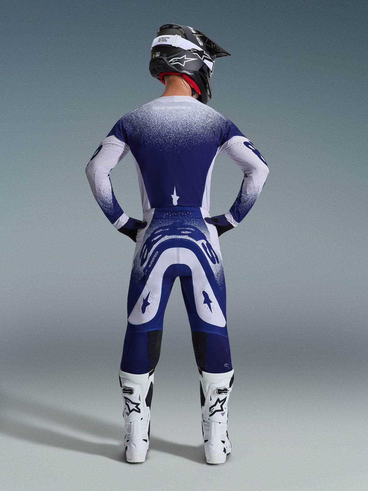 Motocross-Fahrer von hinten im Alpinestars 2026 Supertech Scenz Trikot, Hellgrau und Dunkelblau mit gepunktetem Farbverlauf-Design, passende Motocross-Hose, weiße Rennstiefel und schwarzer Helm, komplette Offroad-Rennausrüstung.