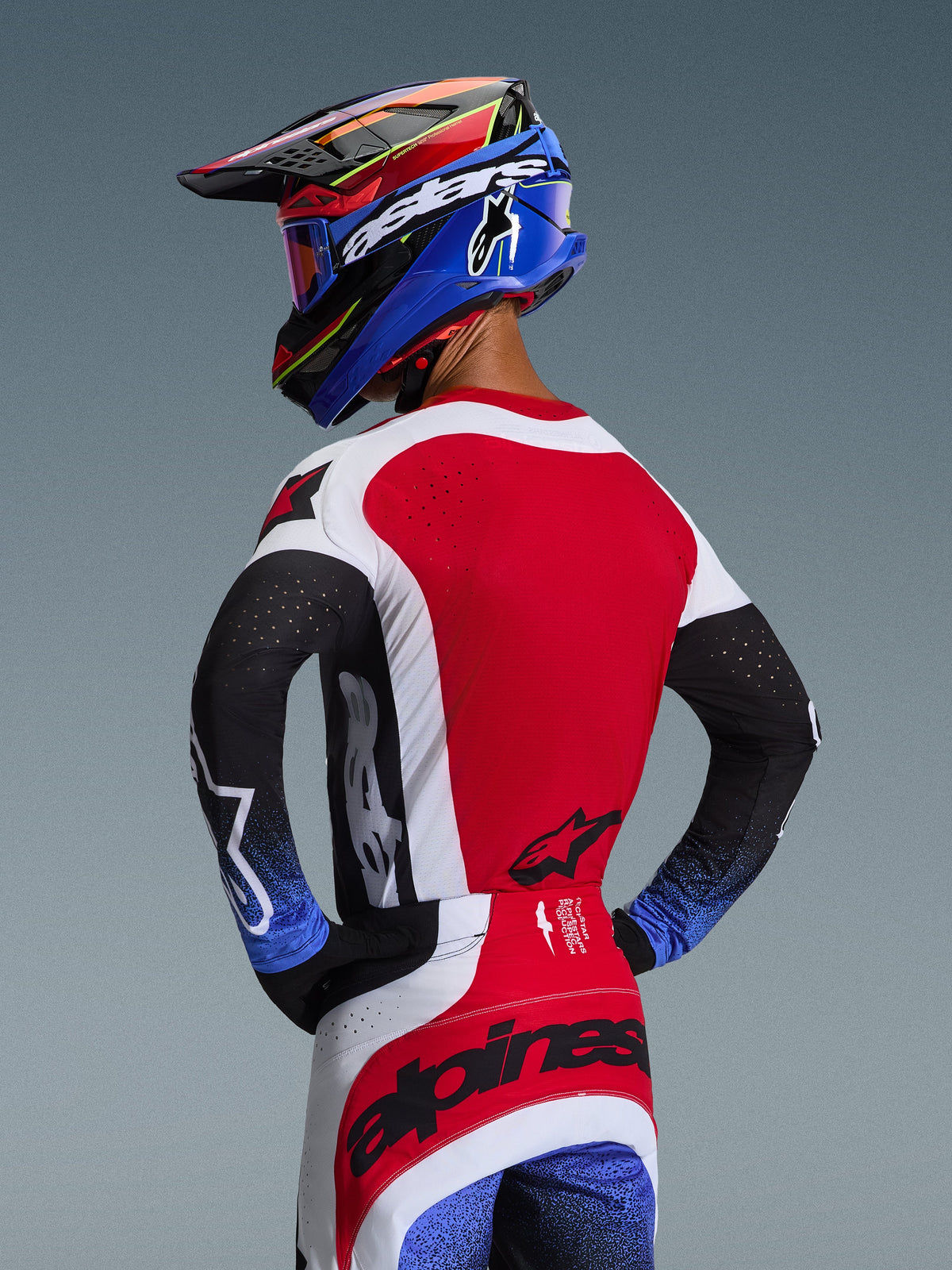 Motocross rider from the back wearing the 2026 Techstar Knif Gear Set, red jersey with white side panels and black sleeves with blue speckled cuffs, red and white pants with black Alpinestars branding across the seat, coordinated blue and red helmet with goggles.