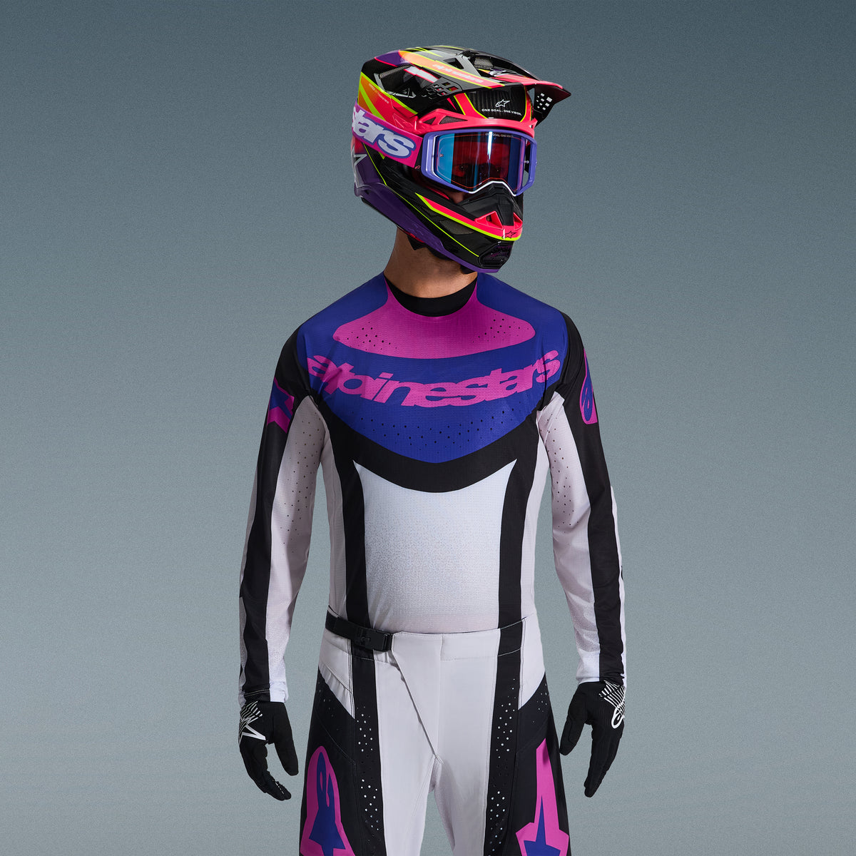 Motocross rider wearing Alpinestars 2026 Techstar Knif Gear Set, Jersey with blue and pink chest graphic and white torso, white pants with black panels and pink logos, multi-colored helmet in purple and pink with matching goggles, black gloves, professional off-road racing gear