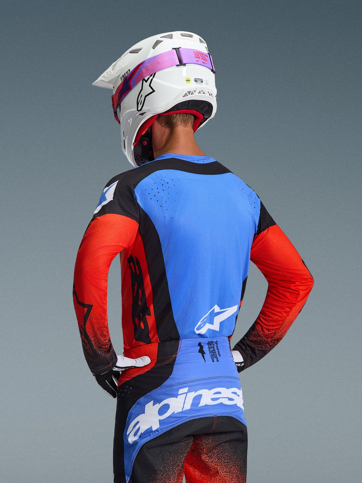 Rear view of a motocross rider wearing the 2026 Techstar Knif Jersey in Orange Black UCLA Blue, featuring a blue back panel with laser-cut perforations, orange sleeves, and black accents, paired with matching motocross pants and a white Alpinestars helmet.