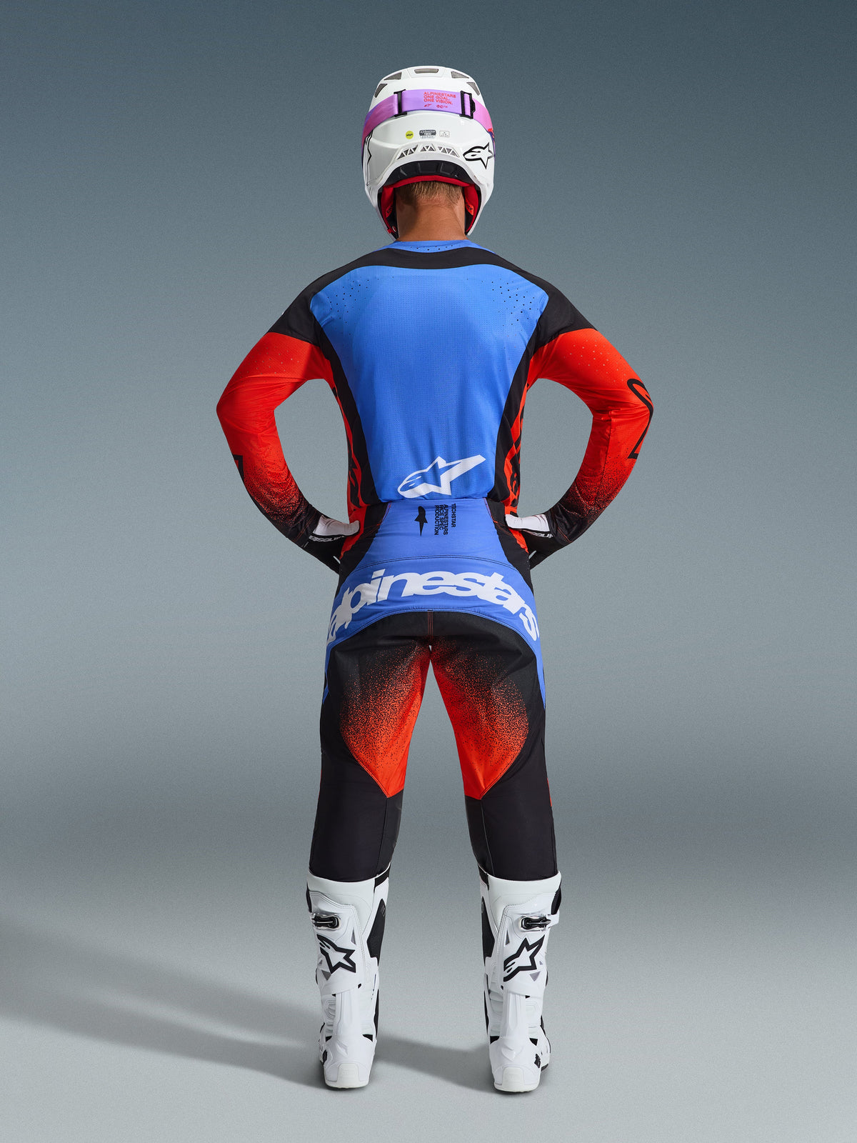 Rückansicht eines Motocross-Fahrers im Alpinestars 2026 Techstar Knif Gear Set, blau-rotes Trikot mit schwarzen Akzenten, schwarze Hose mit rotem Farbverlauf an den Oberschenkeln und blauem Branding, kombiniert mit weißen Rennstiefeln und einem weißen Helm mit pinker Brille.