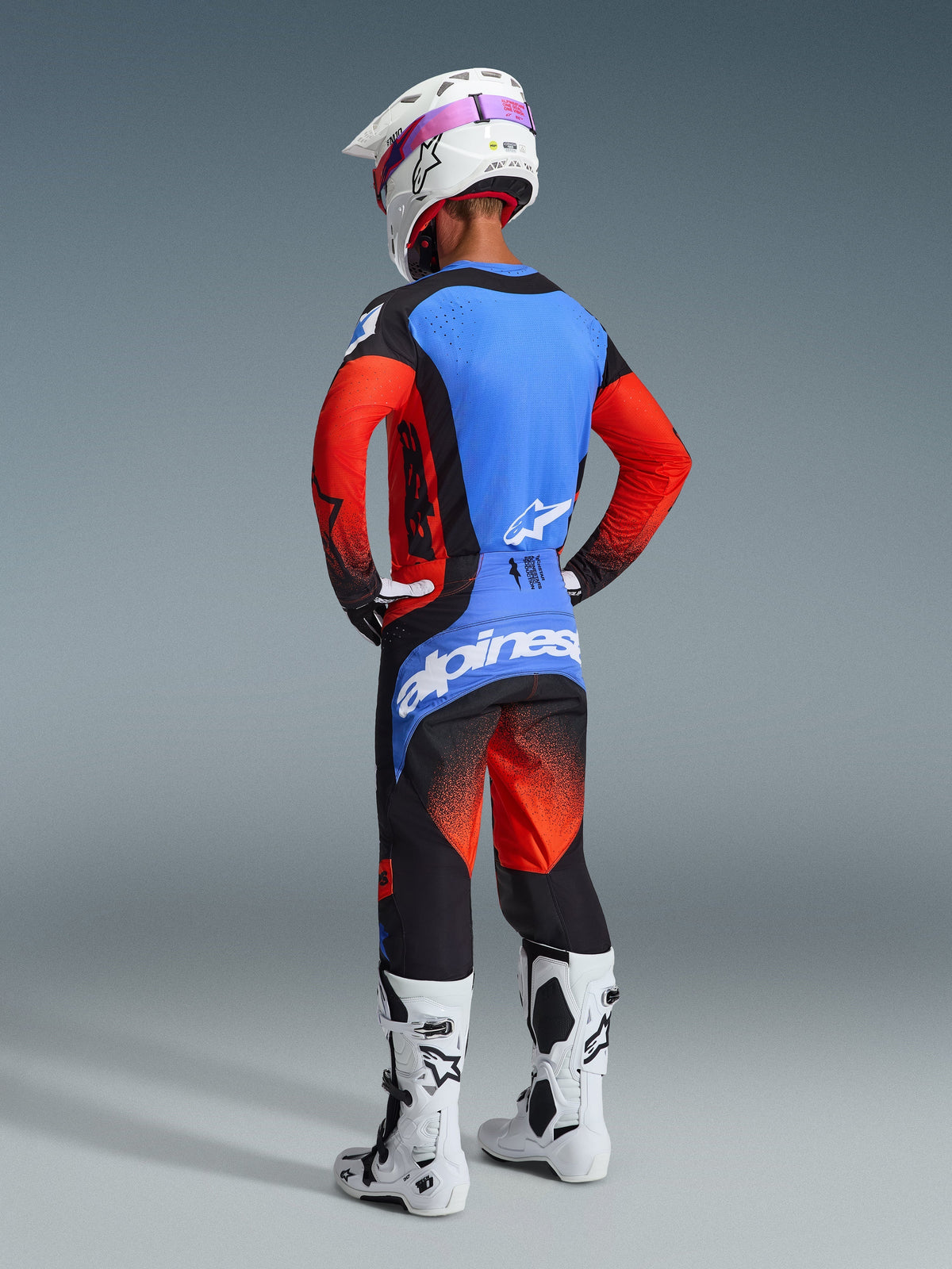 Motocross rider from the back wearing the 2026 Techstar Knif Jersey in Orange, Black, and UCLA Blue, paired with matching motocross pants, a white helmet with pink goggles, and white boots, showing a full Alpinestars off-road racing outfit.