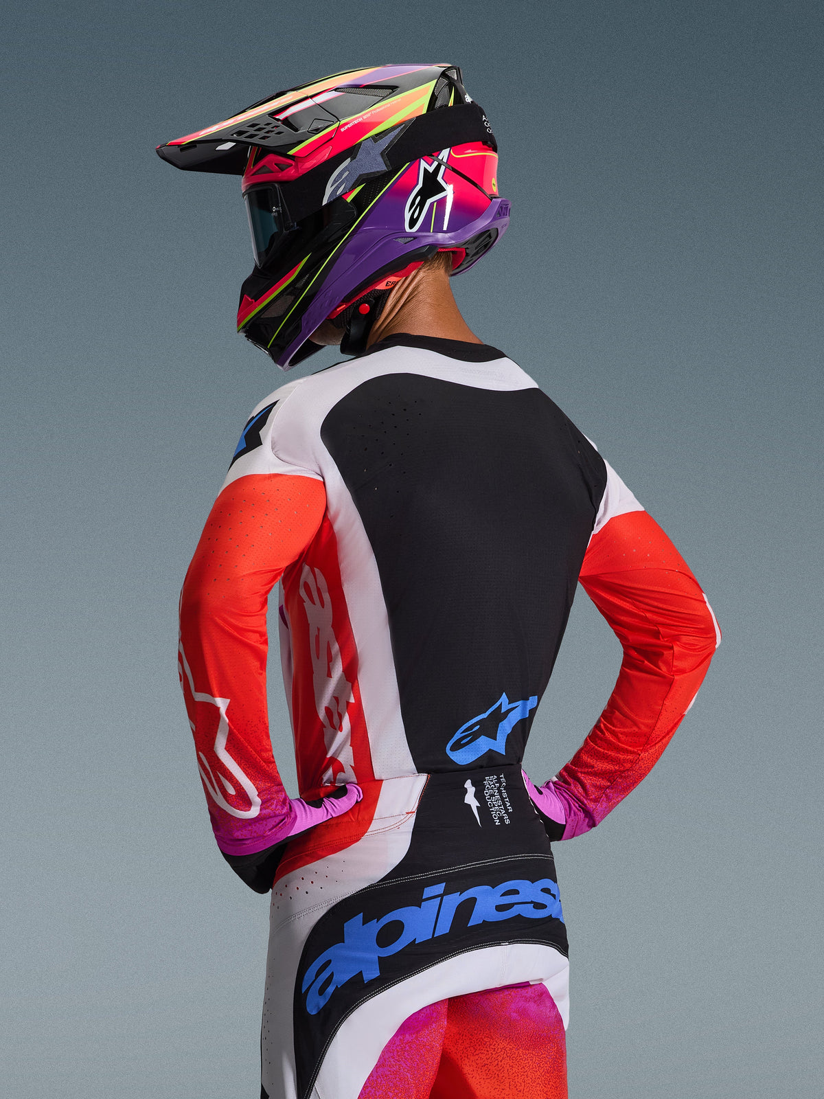 Motocross-Fahrer von hinten im Alpinestars 2026 Techstar Knif Gear Set, neonorangefarbene Ärmel mit schwarzem Rückenteil, weiß-schwarze Hose mit blauen Logos, auffälliger lila und pinker Helm, professionelle Motocross-Ausrüstung für Offroad-Performance