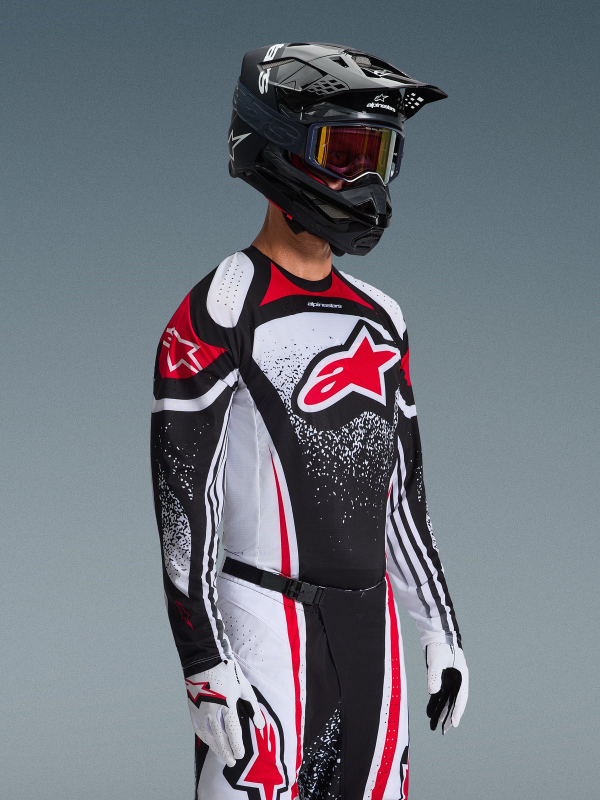 Motocross-Fahrer im Alpinestars 2026 Techstar Nomur Trikot in Schwarz-Weiß-Rot, mit einem großen roten Astars-Logo und gesprenkelten grafischen Mustern, kombiniert mit passender Rennhose, weißen Handschuhen und einem schwarzen Helm mit Goggles, koordiniertes Outfit für den Offroad-Wettbewerb