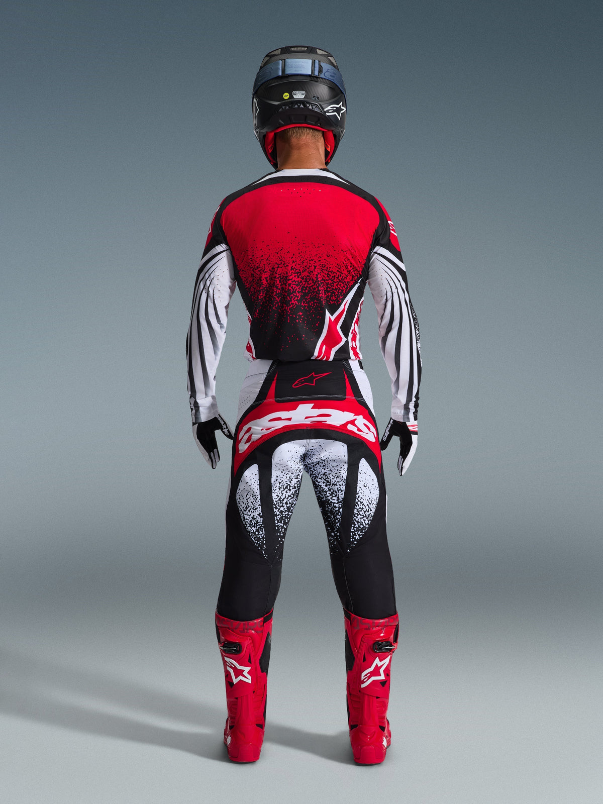 Motocross-Fahrer von hinten im Alpinestars 2026 Techstar Nomur Trikot, Rot, Schwarz und Weiß mit gesprenkeltem Muster, passende Rennhose, rote Motocross-Stiefel und schwarzer Helm, komplette Ausrüstung für Offroad-Wettbewerbe