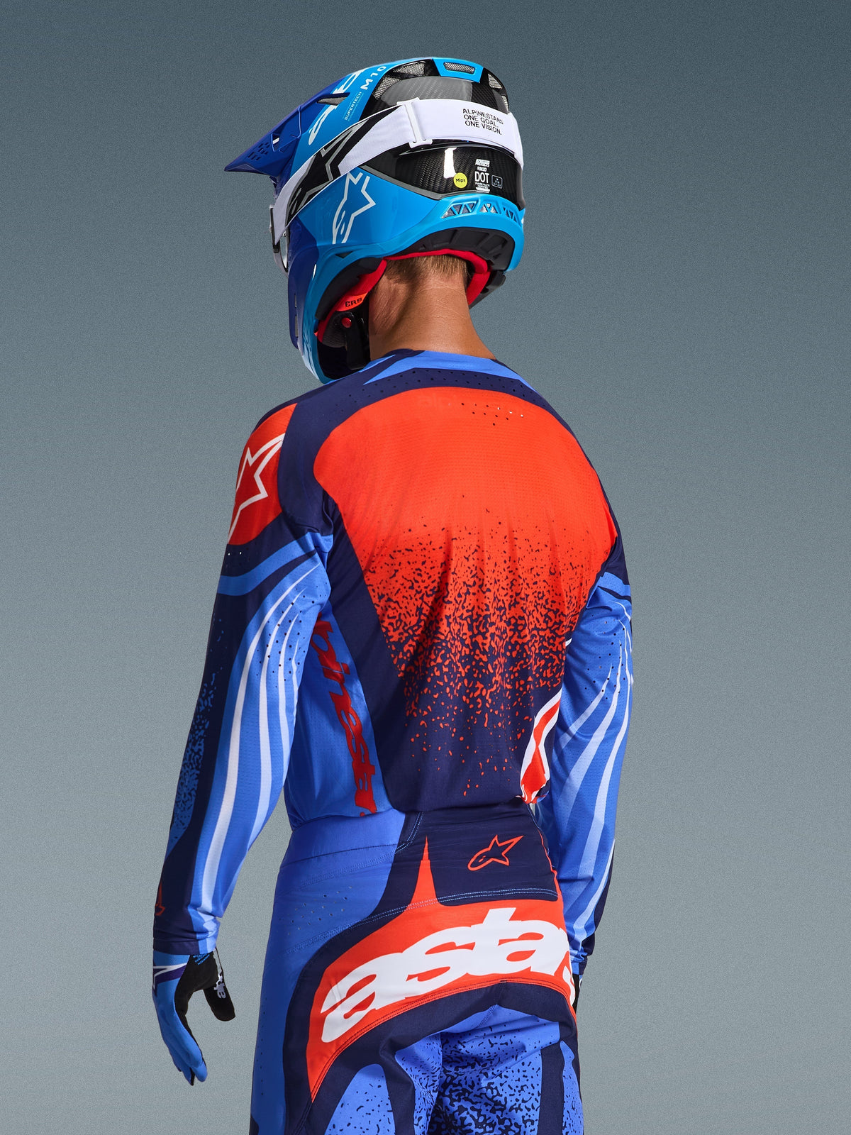 Motocross rider from behind wearing the 2026 Techstar Nomur Jersey, Dark Navy with Orange and UCLA Blue accents, featuring a large orange textured graphic on the back, paired with matching motocross pants and a blue racing helmet, coordinated professional gear for off-road riding.