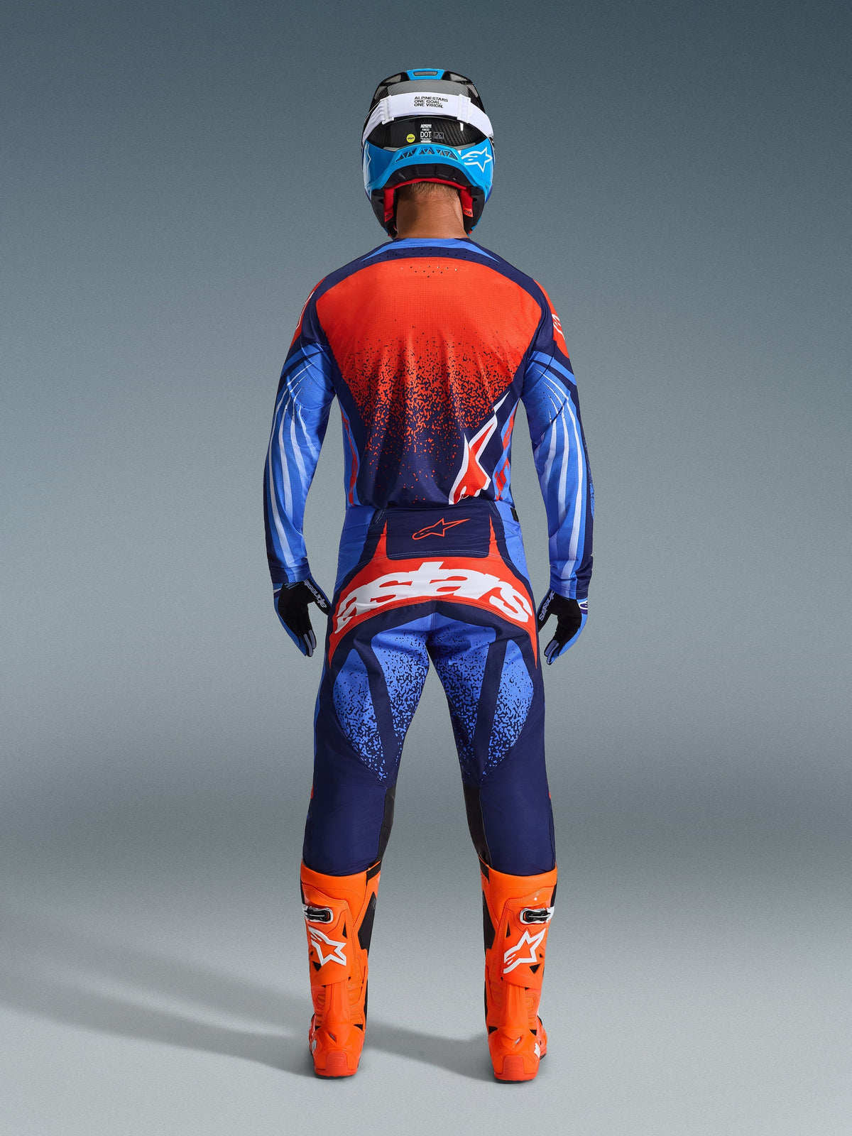 Motocross rider from behind wearing the 2026 Techstar Nomur Jersey and matching pants, Dark Navy and Orange colorway with UCLA Blue accents, bright orange boots, blue and white helmet, and black gloves, complete professional off-road racing gear