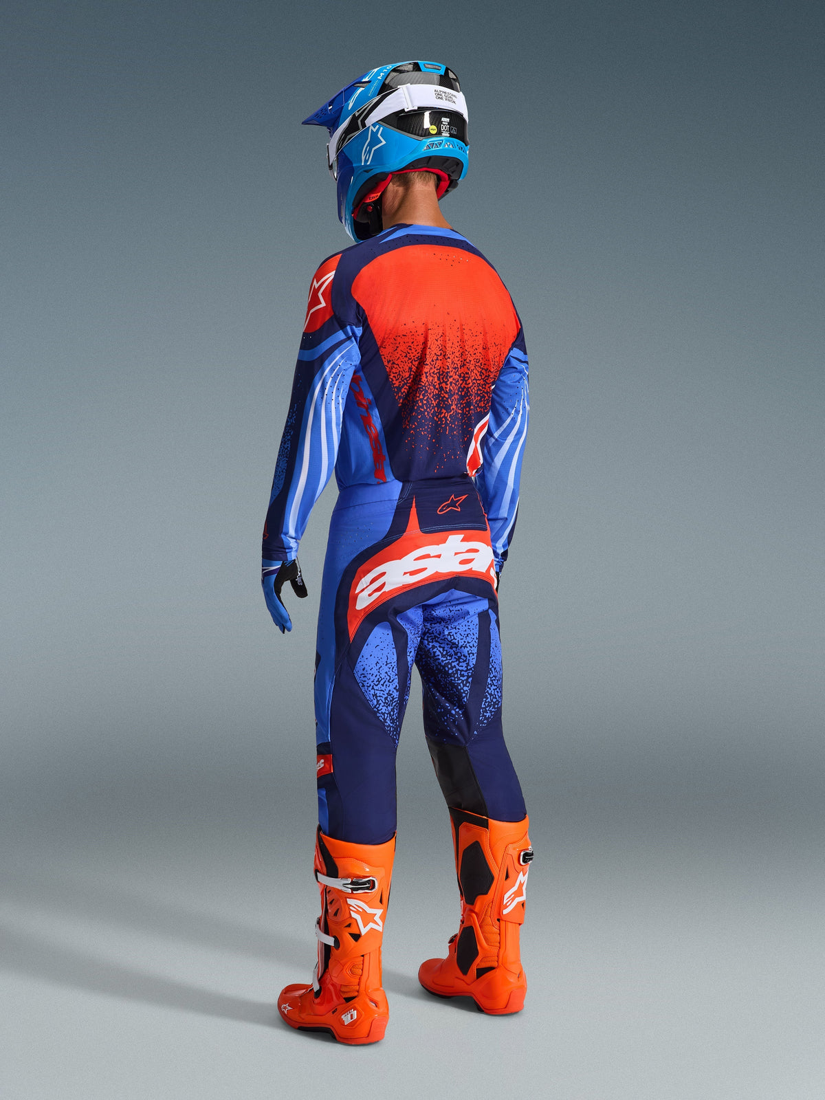 Motocross rider seen from the rear wearing the Alpinestars 2026 Techstar Nomur Jersey in Dark Navy, Orange, and UCLA Blue, paired with matching pants featuring bold white branding, bright orange boots, and a blue and white helmet, complete off-road racing kit.