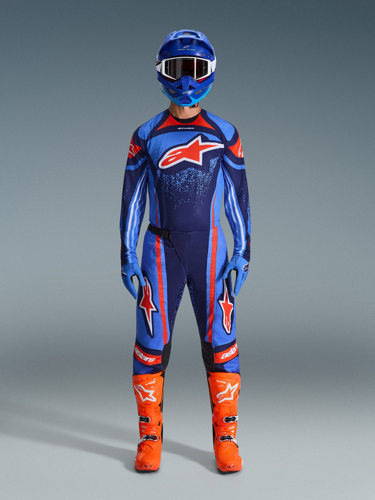 Motocross rider wearing the Alpinestars 2026 Techstar Nomur Jersey in Dark Navy, Orange, and UCLA Blue, paired with matching motocross pants, a blue helmet with goggles, and bright orange racing boots, full coordinated off-road competition gear