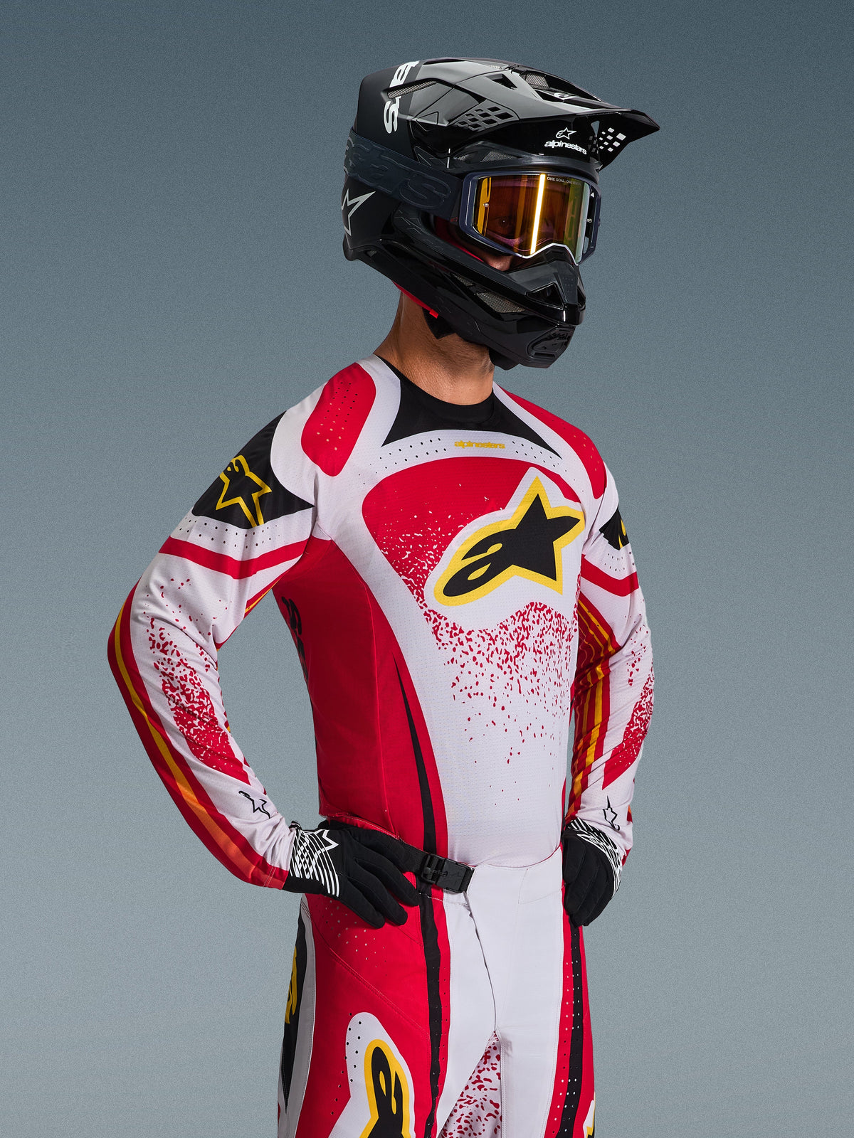 Motocross-Fahrer im Alpinestars 2026 Techstar Nomur Gear Set, weiß-rotes Jersey mit schwarzen Akzenten und gelbem Logo, passende Hose, schwarzer Helm mit gold getönter Brille, schwarz-weiße Handschuhe, athletische Rennpassform für Offroad-Performance