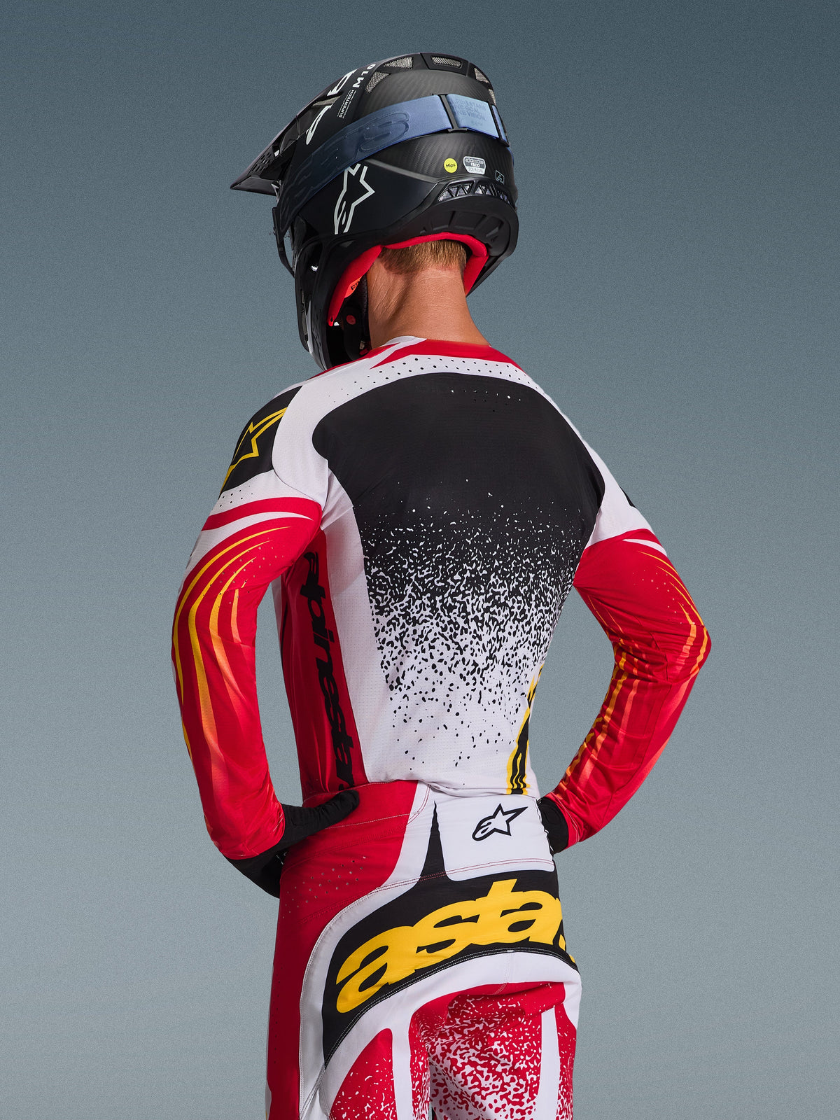 Alpinestars 2026 Techstar Nomur Gear Set, Motocross-Trikot und -Hose, Rot, Weiß und Schwarz mit gelben Akzenten, Rückansicht eines Fahrers mit schwarzem Helm, Performance-Rennbekleidung mit Splatter-Grafiken und markantem Branding