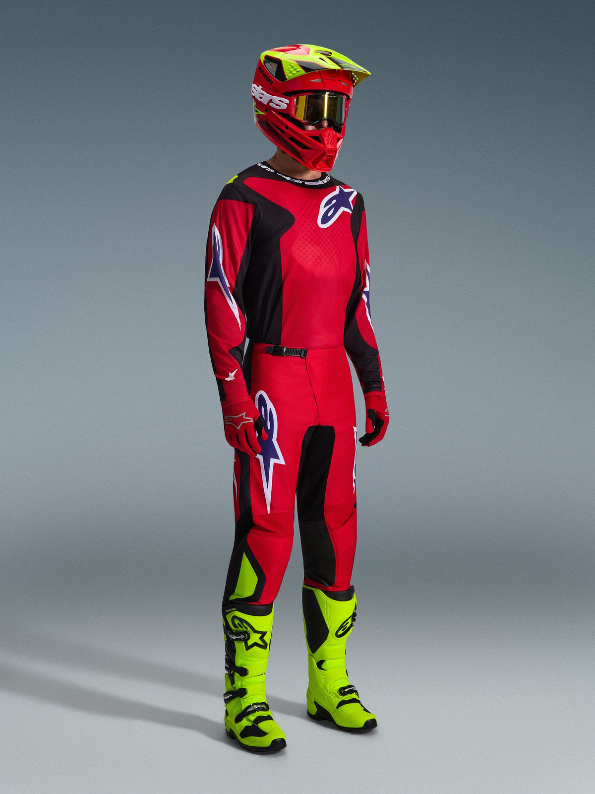 Motocross-Fahrer im Alpinestars 2026 Fluid Grid Gear Set in Rot, Schwarz und Lila, mit passendem Trikot und Hose mit lila Logos, einem rot-neongelben Helm und neongelben Stiefeln für eine komplette Offroad-Rennausrüstung.