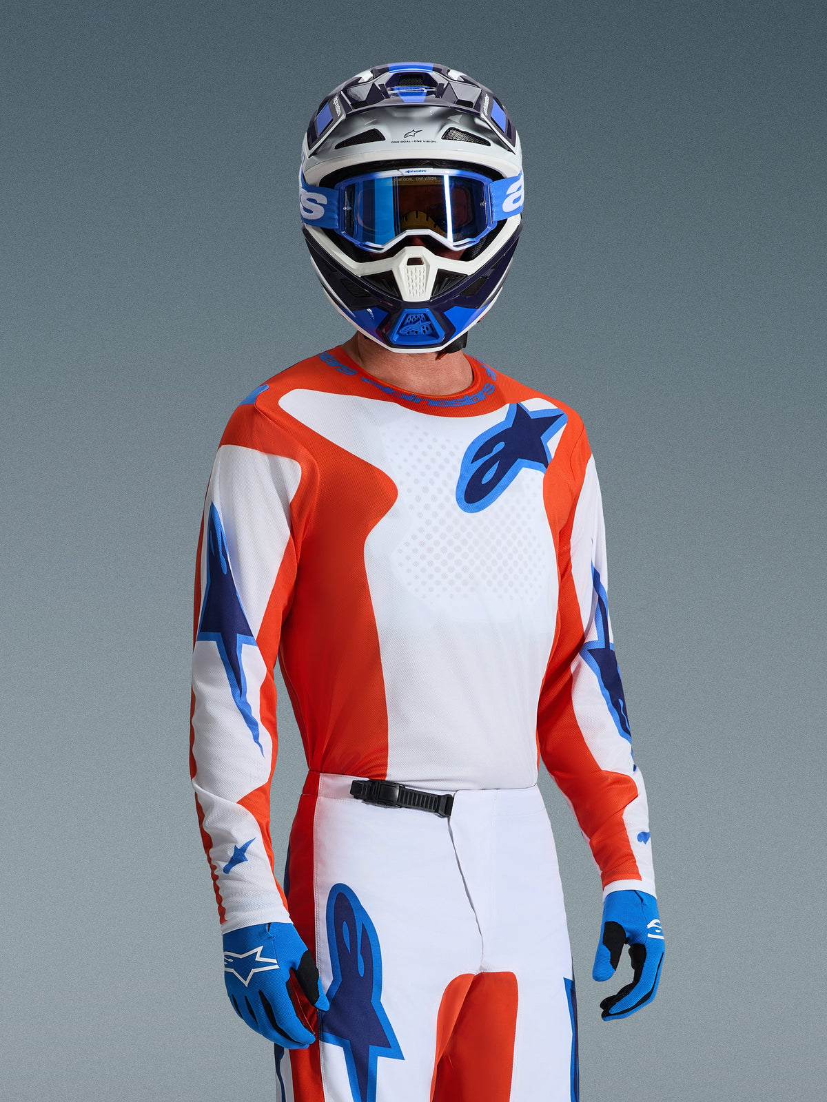 Motocross-Fahrer trägt das Alpinestars 2026 Fluid Grid Ausrüstungsset, orange-weißes Trikot mit blauen Stern-Logos, weiß-orangefarbene Hose mit blauen Akzenten, blaue Handschuhe, blau-weißer Helm mit blauer Brille, abgestimmte sportliche Rennausrüstung für Offroad-Performance