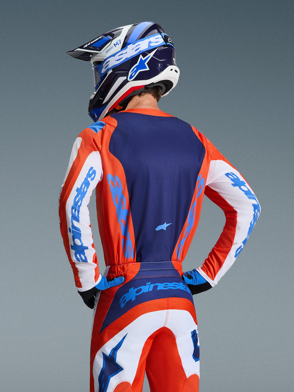 Alpinestars 2026 Fluid Grid Trikot, Motocross-Trikot, Orange und UCLA-Blau, Rückansicht eines Fahrers in passender Hose und Helm, mit dunkelblauem Rückenteil und weißen Ärmeln mit hellblauem Branding, sportliche Passform für Offroad-Rennen