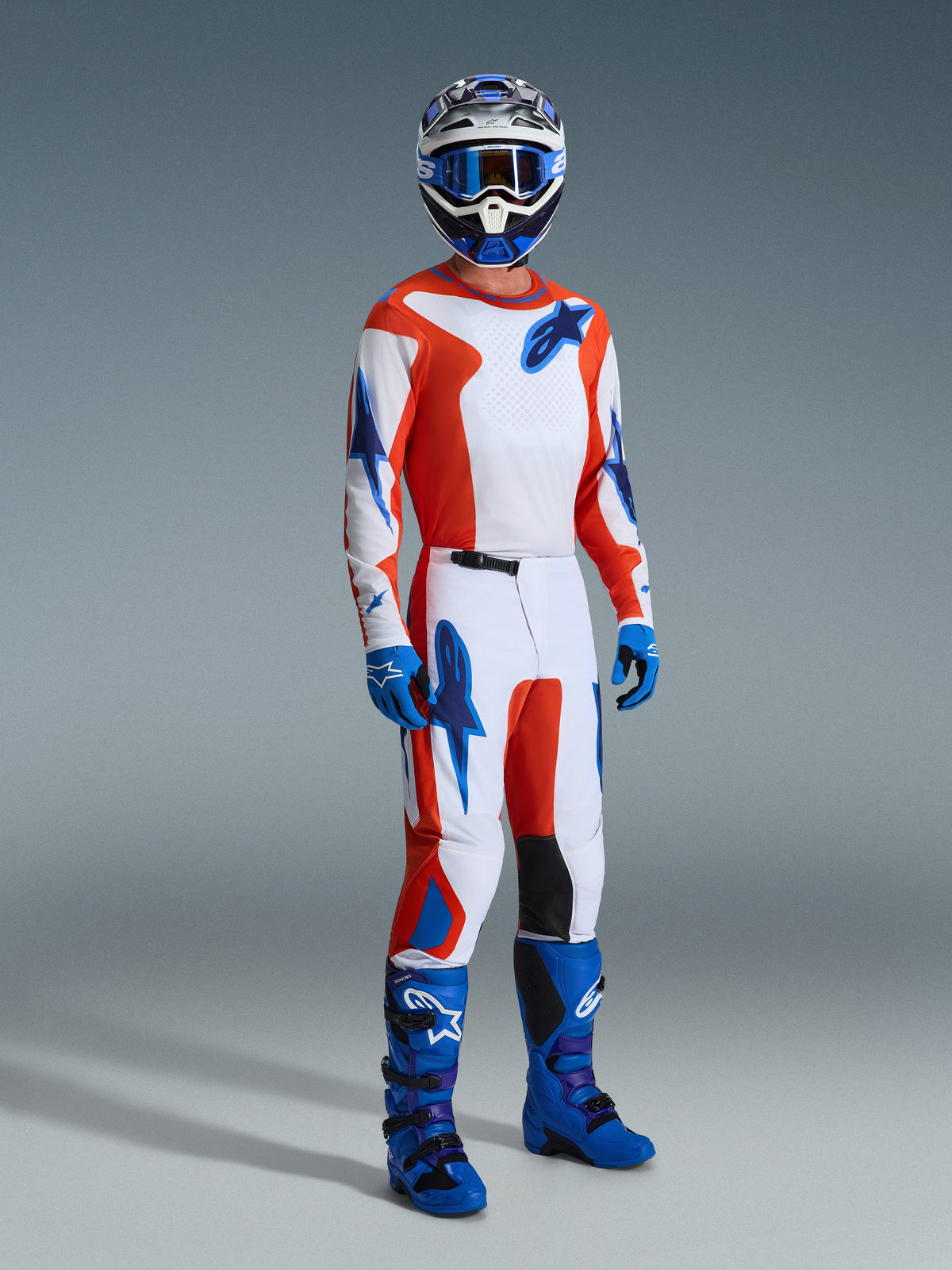 Motocross-Fahrer im Alpinestars 2026 Fluid Grid Gear Set, weißes Trikot und Hose mit orangefarbenen und UCLA-blauen Grafiken, passender blauer Helm und Brille, blaue Handschuhe und leuchtend blaue Rennstiefel, koordiniertes Performance-Outfit für das Gelände.
