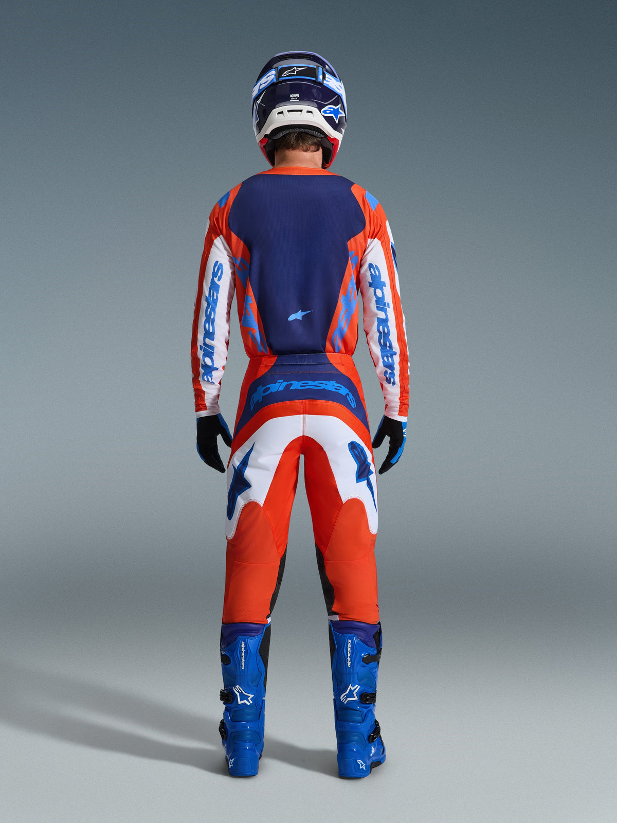 Alpinestars 2026 Fluid Grid Bekleidungsset, Motocross-Fahrer in der Rückansicht in einem kompletten Rennoutfit in Orange und UCLA Blue, langärmliges Trikot mit marineblauem Rücken und weißen Ärmeln, passender orangefarbener Hose mit blauen Grafiken, blauen Stiefeln und abgestimmtem Helm, professionelle Offroad-Rennausrüstung.