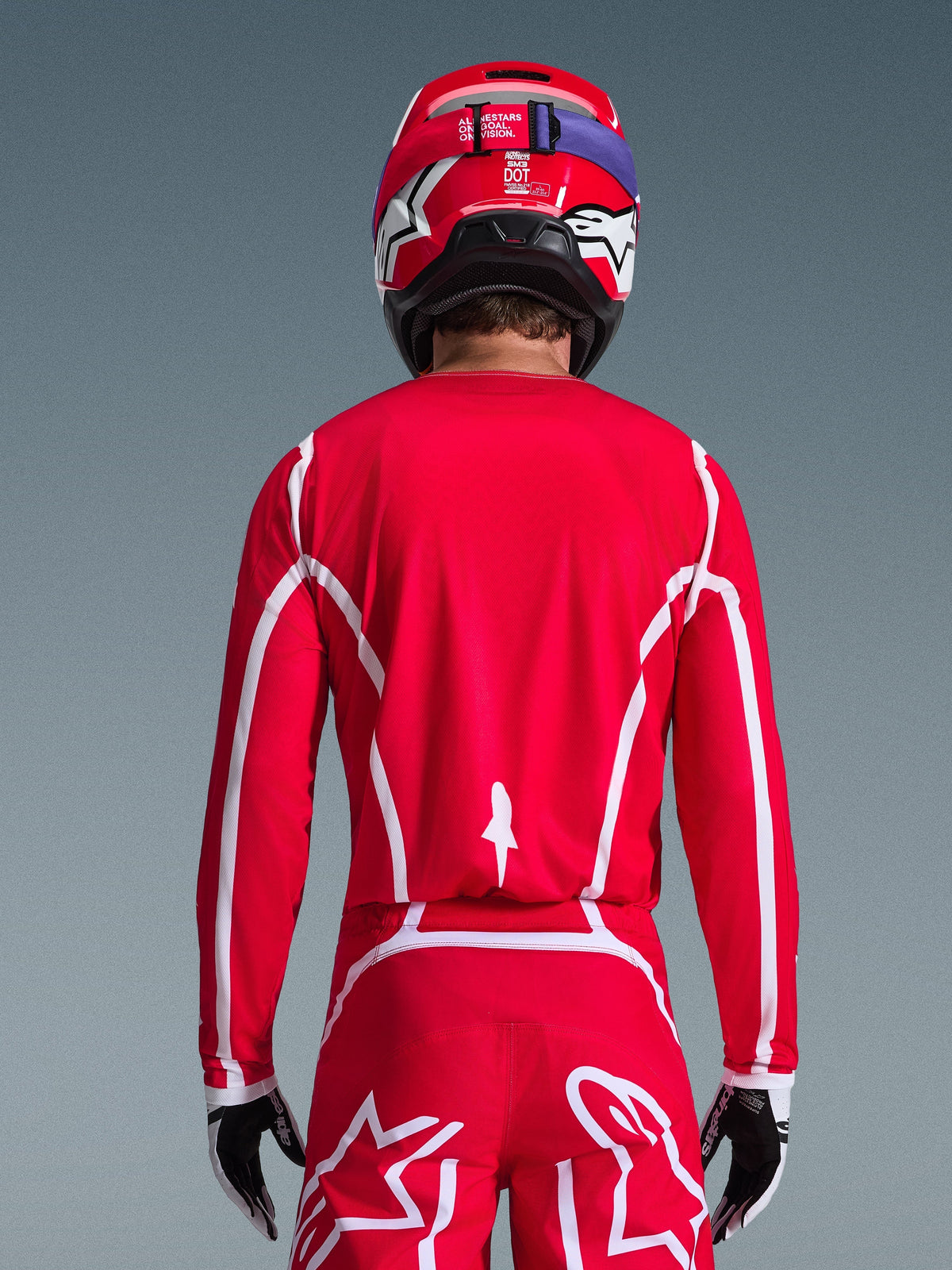 Rückansicht eines Motocross-Fahrers im Alpinestars 2026 Fluid Apex Trikot in Rot und Weiß, mit geometrischen weißen Mustern, kombiniert mit passender Motocross-Hose und einem roten Helm, komplette Rennausrüstung für Offroad-Performance.