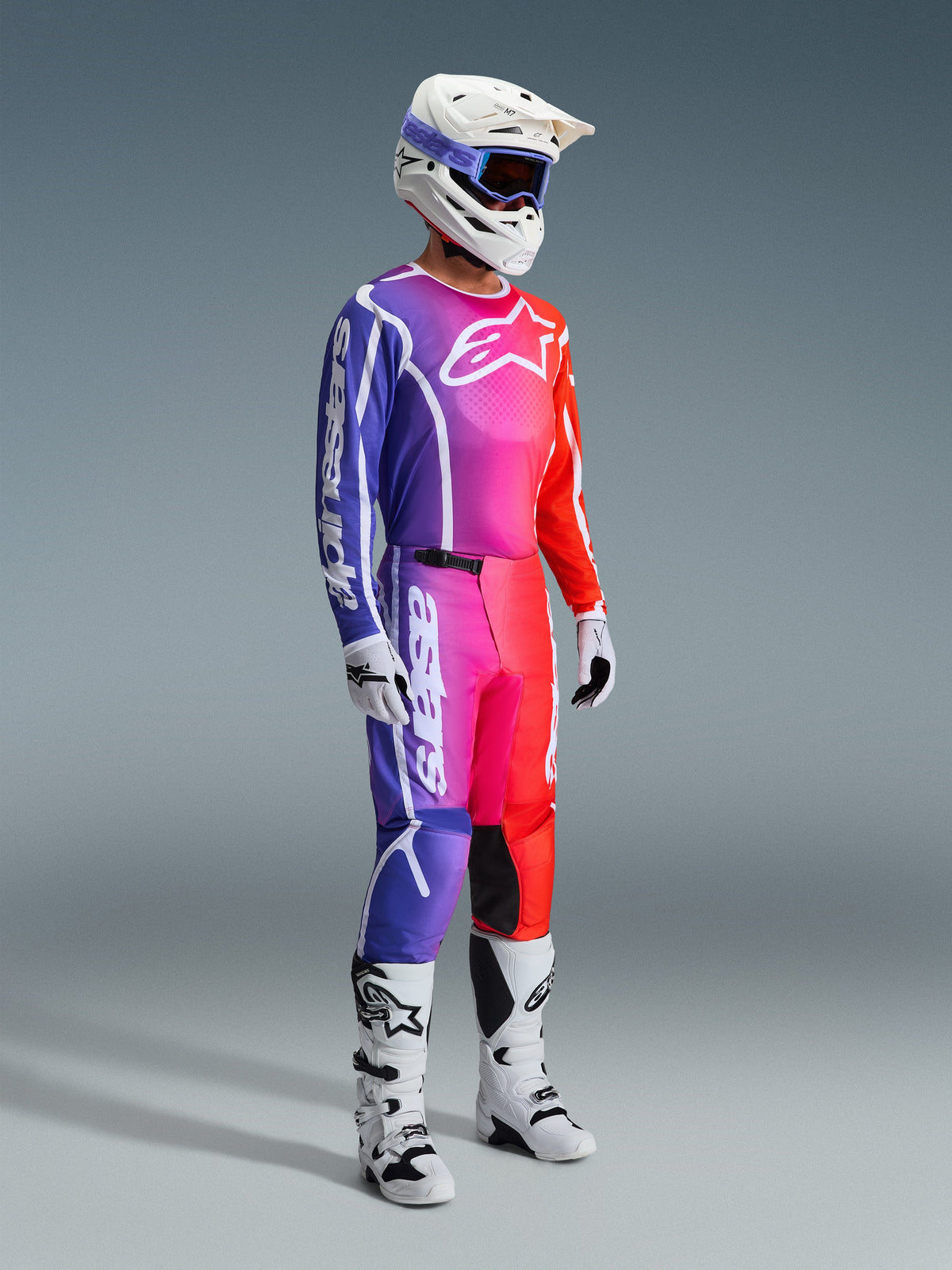 Motocross-Fahrer in kompletter Rennausrüstung, 2026 Fluid Apex Trikot und Hose mit einem lebendigen Farbverlauf in Lila, Pink und Orange, weißer Helm mit blauer Brille, weiß-schwarze Stiefel und weiße Handschuhe, abgestimmtes Outfit für Offroad-Motocross.