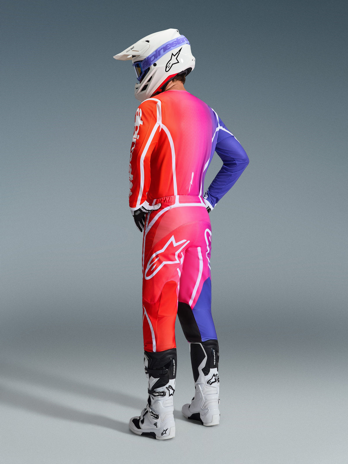 Motocross-Fahrer im Alpinestars 2026 Fluid Apex Trikot und passender Hose, leuchtender Farbverlauf in Rot, Pink und Blau mit weißen Akzenten, weißer Helm mit blauer Brille und weiße Stiefel, Rückansicht der kompletten Offroad-Rennausrüstung.