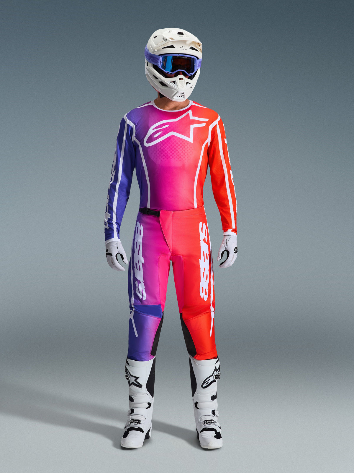 Motocross-Fahrer in kompletter Alpinestars 2026 Fluid Apex Rennausrüstung, mit Trikot und Hose in einem lila, pinken und orangefarbenen Farbverlauf. Das abgestimmte Outfit umfasst ein weißes Alpinestars-Logo auf der Brust, einen weißen Helm mit blauer Brille und weiße Stiefel, entwickelt für Offroad-Performance.