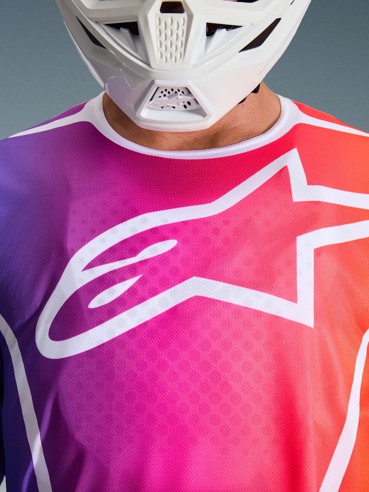 Alpinestars 2026 Fluid Apex Jersey, Motocross Jersey, Purple Pink and Orange gradient, large white Alpinestars star logo on chest, close-up view showing white helmet chin guard