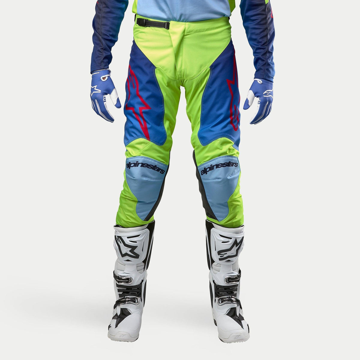 Alpinestars 2024 Racer Hoen Hose, Motocross-Hose, Gelb Fluo und Blau Night Navy, getragen mit weiß-schwarzen Motocross-Stiefeln und blauen Handschuhen, mit markanten roten Logos, hellblauen Knieeinsätzen und einem sportlichen ergonomischen Design für den Offroad-Einsatz.