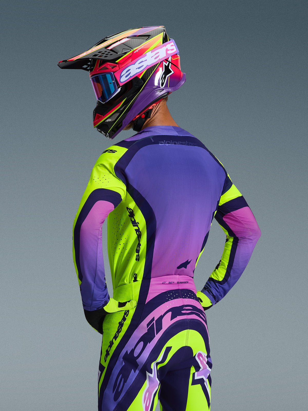 Motocross rider seen from behind wearing the 2026 Alpinestars Supertech Vista Jersey and matching pants, Yellow Purple Black colorway with vibrant gradients, coordinated racing helmet with blue goggles and yellow gloves, professional off-road racing kit showcasing an athletic fit and integrated branding.