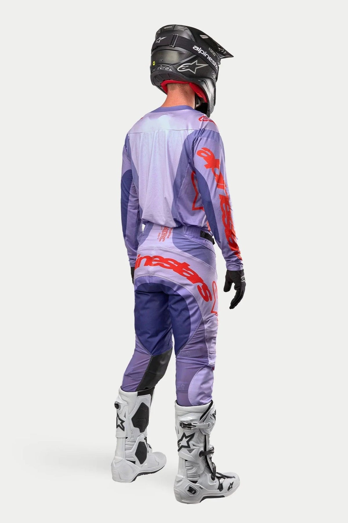 Motocross-Fahrer in kompletter Rennausrüstung von hinten, lila Alpinestars 2024 Techstar Ocuri Trikot mit leuchtend orangefarbenem Branding, passende lila Hose mit orangefarbenen Details, schwarzer Helm, weiß-schwarze Stiefel, schwarze Handschuhe, professionelle Offroad-Rennausrüstung