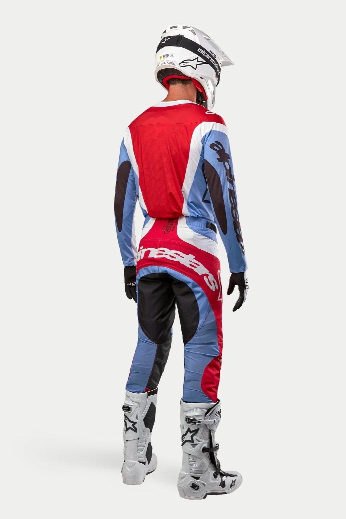 Motocross-Fahrer von hinten gesehen in kompletter Rennausrüstung, 2024 Techstar Ocuri Jersey in Hellblau, Mars-Rot und Weiß, passende Hose mit markantem Branding, weiß-schwarze Stiefel, weißer Helm, abgestimmtes Outfit für den Offroad-Rennsport
