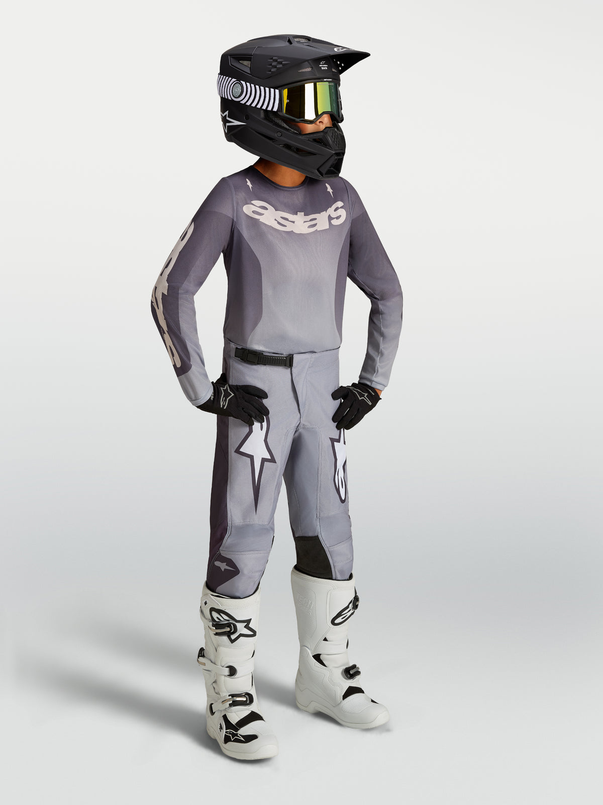 Alpinestars Jugend Fluid Amber Gear Set, graues Motocross-Trikot und -Hose mit weißen Rennstiefeln und schwarzem Helm.