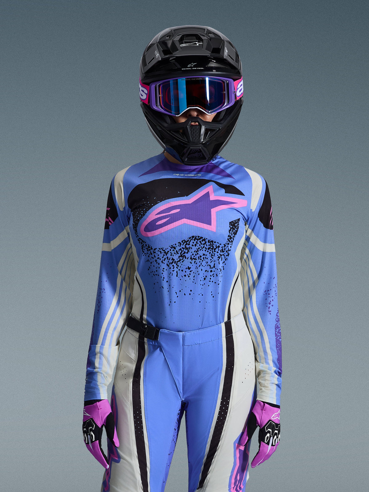 Female motocross rider wearing the Alpinestars 2026 Stella Techstar Nomur Gear Set, blue jersey featuring a pink and purple star logo, matching blue pants with white and black side panels, black helmet with purple and blue goggles, and pink and black gloves, complete women's off-road racing kit