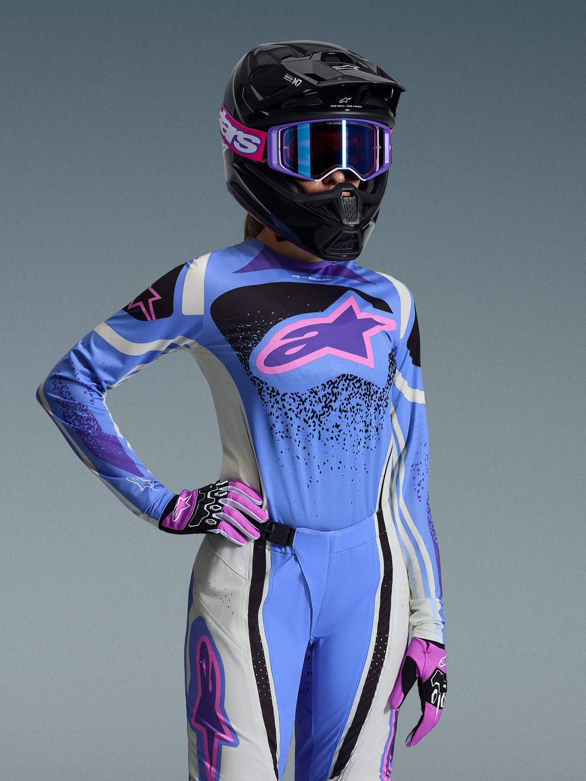 Female motocross rider wearing the Alpinestars 2026 Stella Techstar Nomur Gear Set, light blue jersey with pink and purple accents, matching blue and white pants, black full-face helmet with purple goggles, and pink and black gloves, professional off-road racing gear for women.