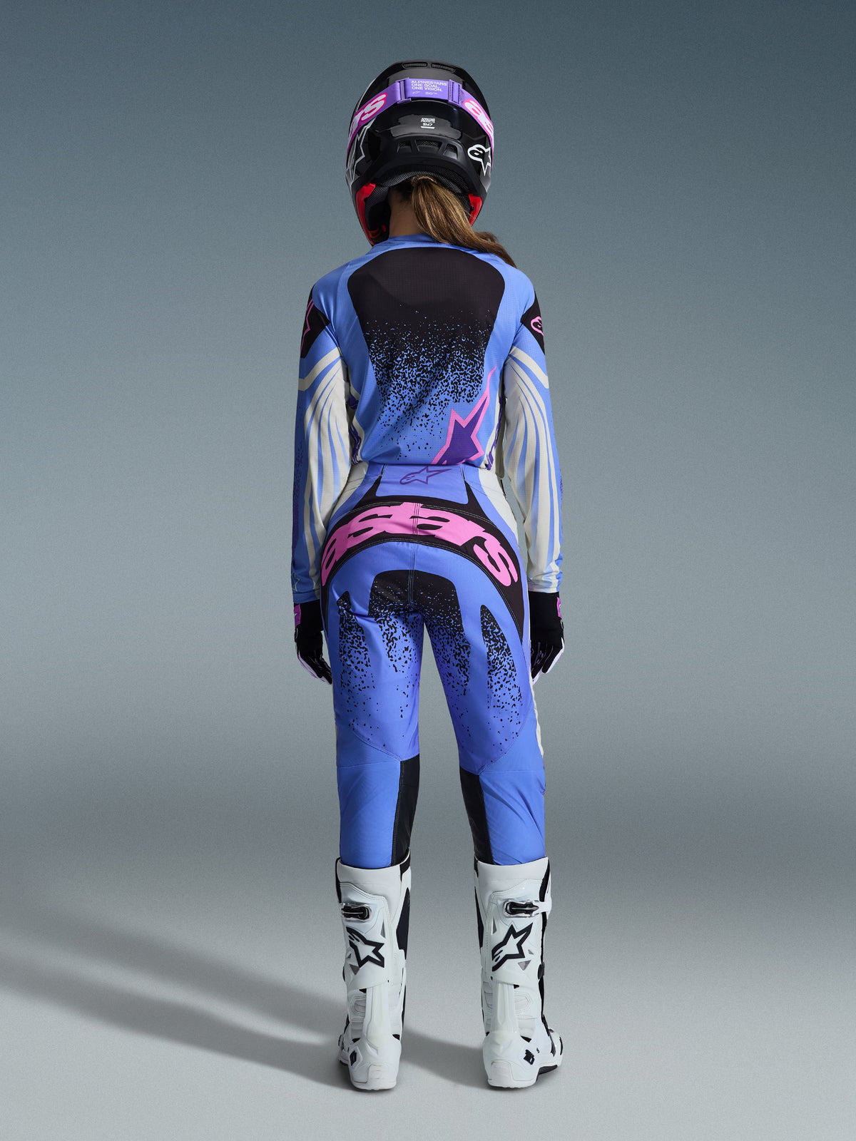 Rear view of a motocross rider wearing the Alpinestars 2026 Stella Techstar Nomur Gear Set, blue jersey and pants with pink and white accents, bold pink branding on the back of the pants, paired with white motocross boots and a coordinated black and pink helmet.