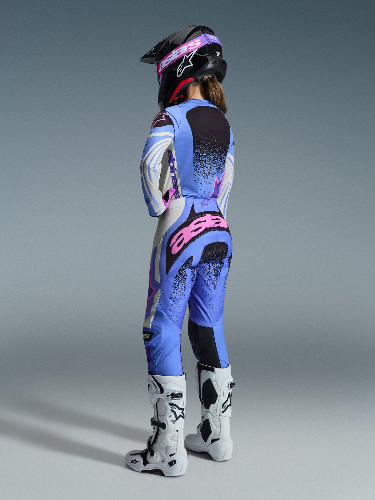 Alpinestars 2026 Stella Techstar Nomur Gear Set, rear view of a female motocross rider wearing a light blue, white, and pink jersey and pants set with black speckled details, paired with a black helmet and white boots, professional racing gear for women.