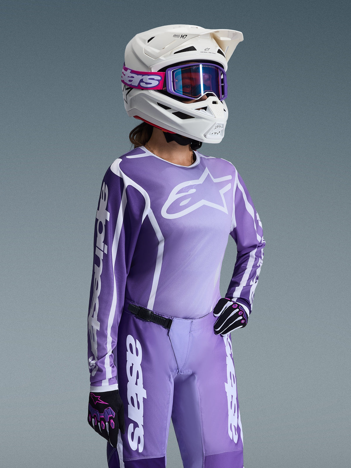 Motocross-Fahrerin im Alpinestars 2026 Stella Fluid Apex Bekleidungsset für Damen, lila-weißes Trikot mit Alpinestars-Logos, passende lila Hose, weißer Helm mit lila Brille und schwarz-lila Handschuhe, professionelle Offroad-Rennausrüstung