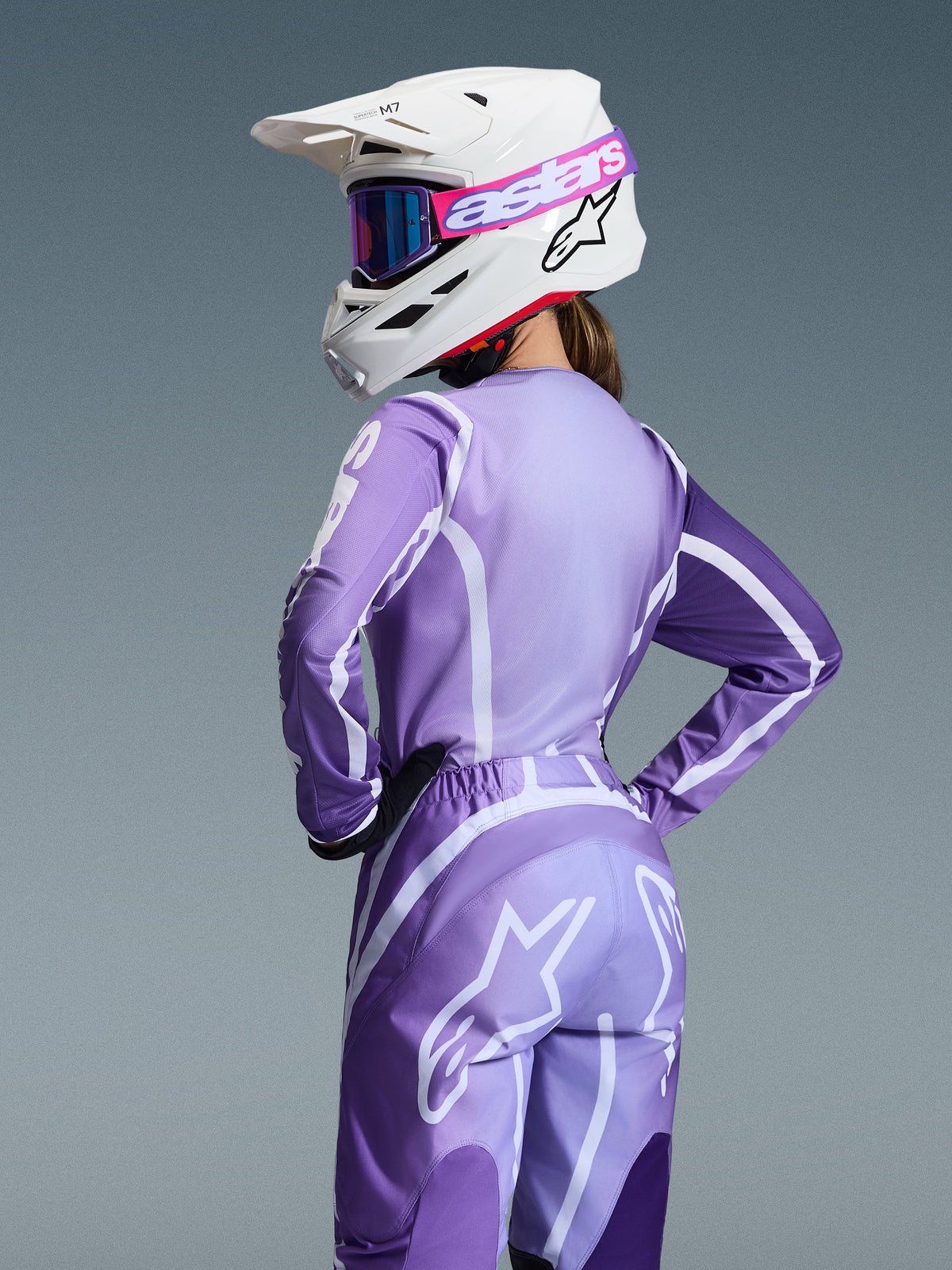 Motocross-Fahrerin im Alpinestars 2026 Stella Fluid Apex Gear Set, lila und lavendelfarbenes Trikot und Hose mit weißen Grafiken, weißer Helm mit pinkfarbener und lila Brille, spezielle Damenpassform, abgestimmte Ausrüstung für den Offroad-Rennsport