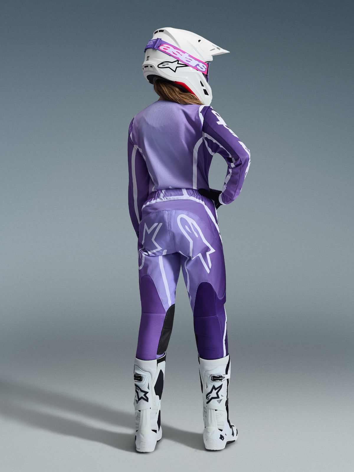 Alpinestars 2026 Stella Fluid Apex Gear Set, Damen-Motocross-Ausrüstung, Lila und Lavendel mit weißen Akzenten, Fahrerin von hinten in passendem Trikot und Hose, weißer Helm mit pinker Brille, weiße Stiefel, koordiniertes sportliches Rennoutfit