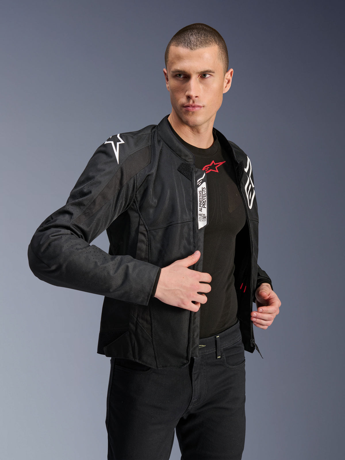 Alpinestars Viper Air V4 Jacke, schwarze Textil-Motorradjacke, getragen von männlichem Model mit schwarzen Jeans und Baselayer, geöffneter Frontreißverschluss zeigt Tech-Air Ready und Alpinestars Protects Labels, weiße Logos auf den Schultern, Mesh-Einsätze für Luftzirkulation